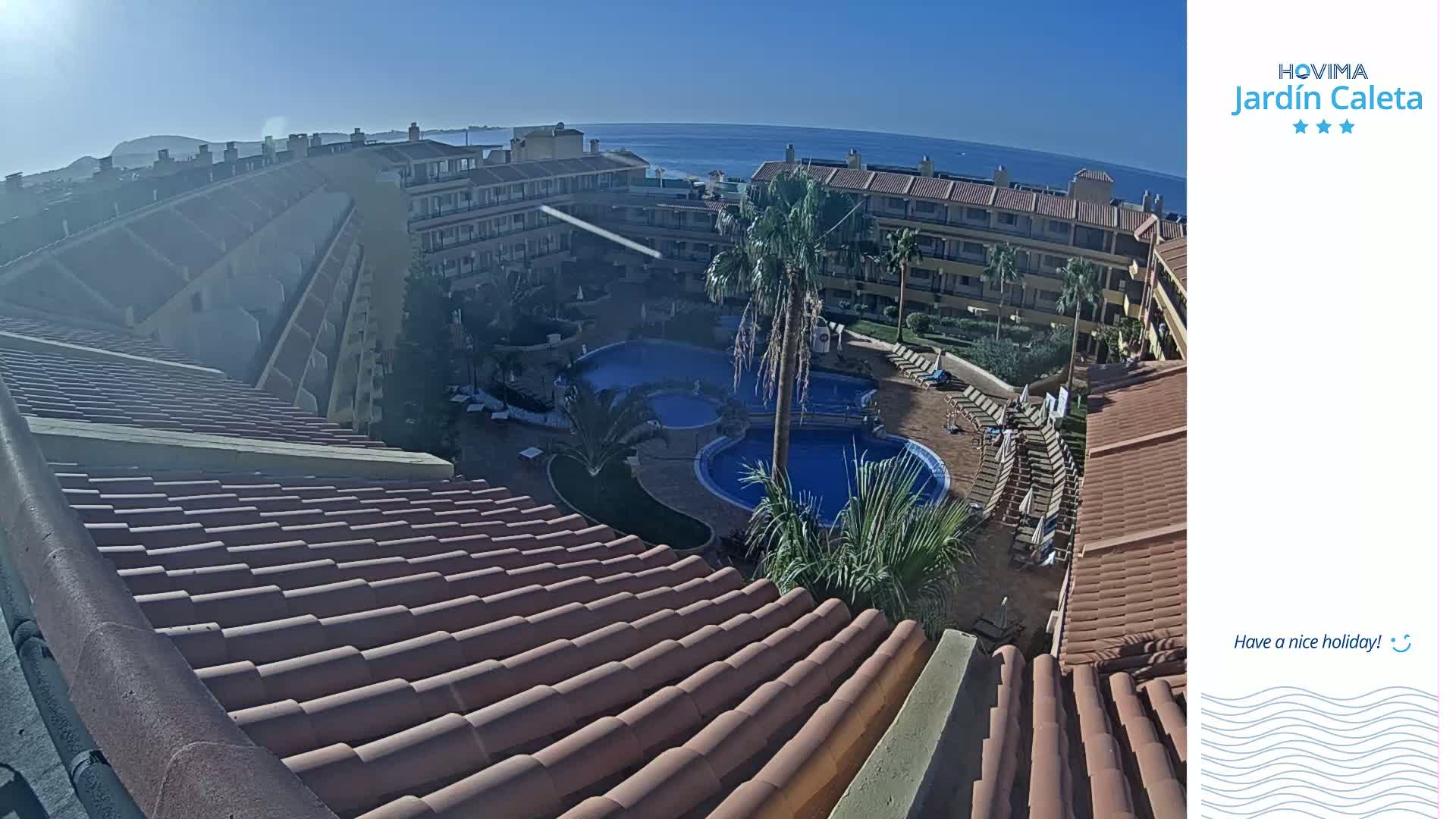 A sunny day overlooks a resort with multiple buildings surrounding a courtyard containing several swimming pools and lounge chairs.