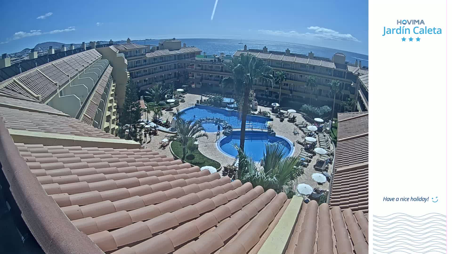 A tiled roof overlooks a courtyard with two swimming pools, lounge chairs, and palm trees, all under a sunny sky and with an ocean view in the distance.
