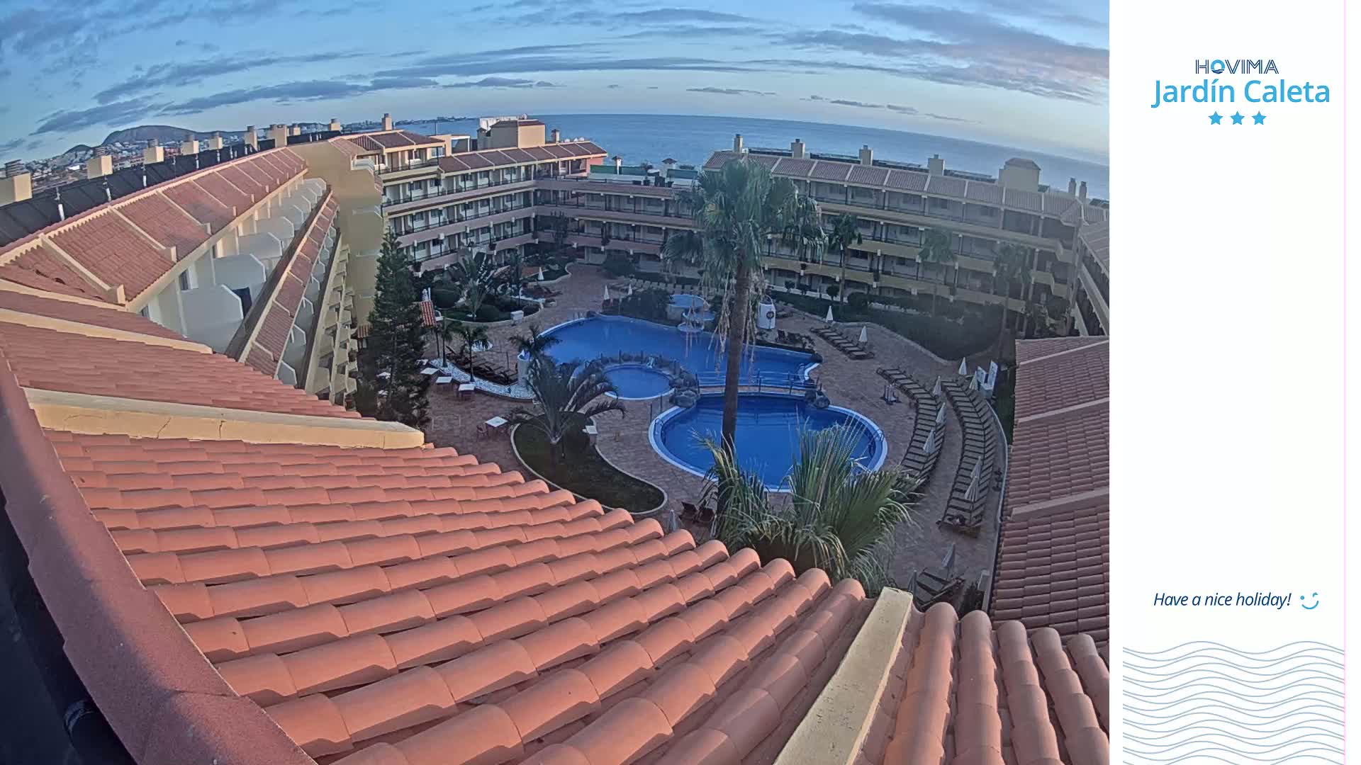 A red-tiled roof overlooks a resort courtyard with a swimming pool complex, palm trees, and an ocean view under a clear, twilight sky.