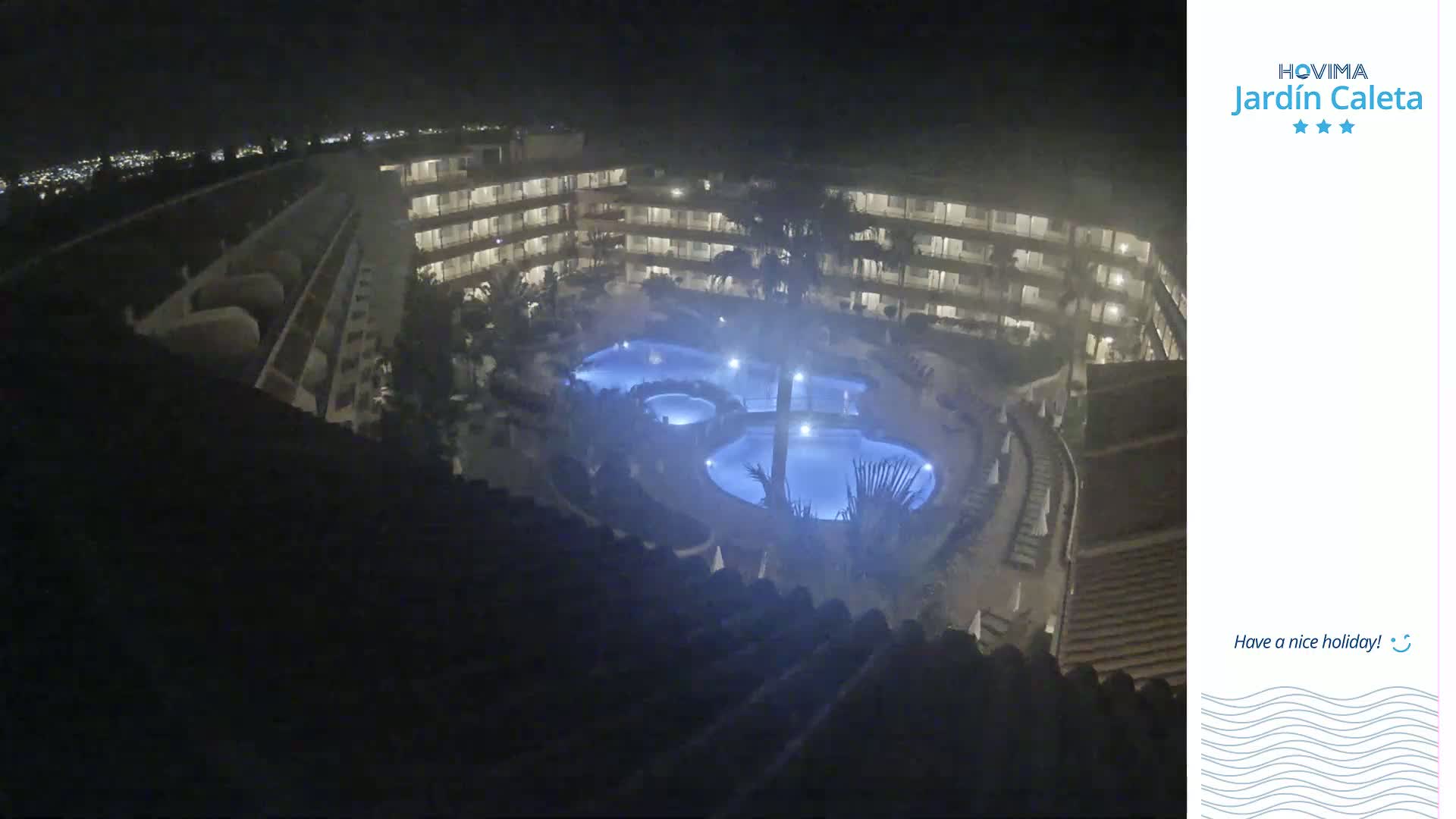 A nighttime, high-angle view shows a multi-story building complex surrounding a lit, multi-section swimming pool.