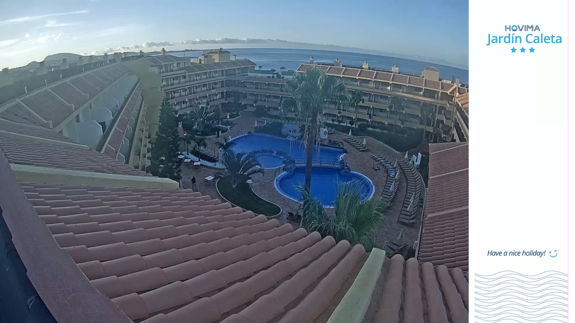 An aerial view shows a resort with multiple buildings surrounding a large swimming pool complex on a clear day with a calm ocean in the background.
