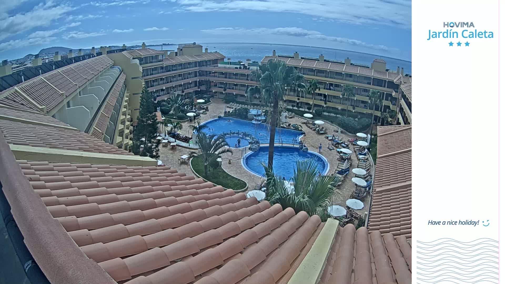 An overhead view of a resort complex with multiple buildings surrounding a courtyard containing two swimming pools on a partly cloudy day.