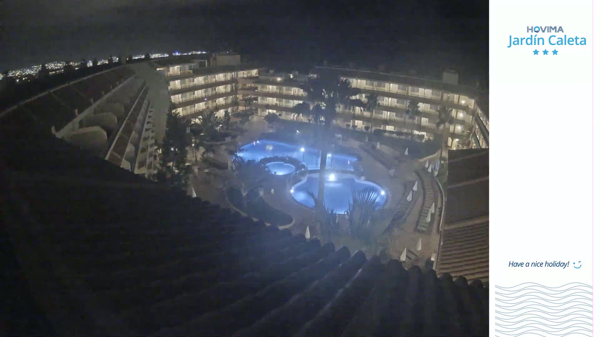 A nighttime aerial view shows a multi-story hotel complex surrounding several illuminated swimming pools.