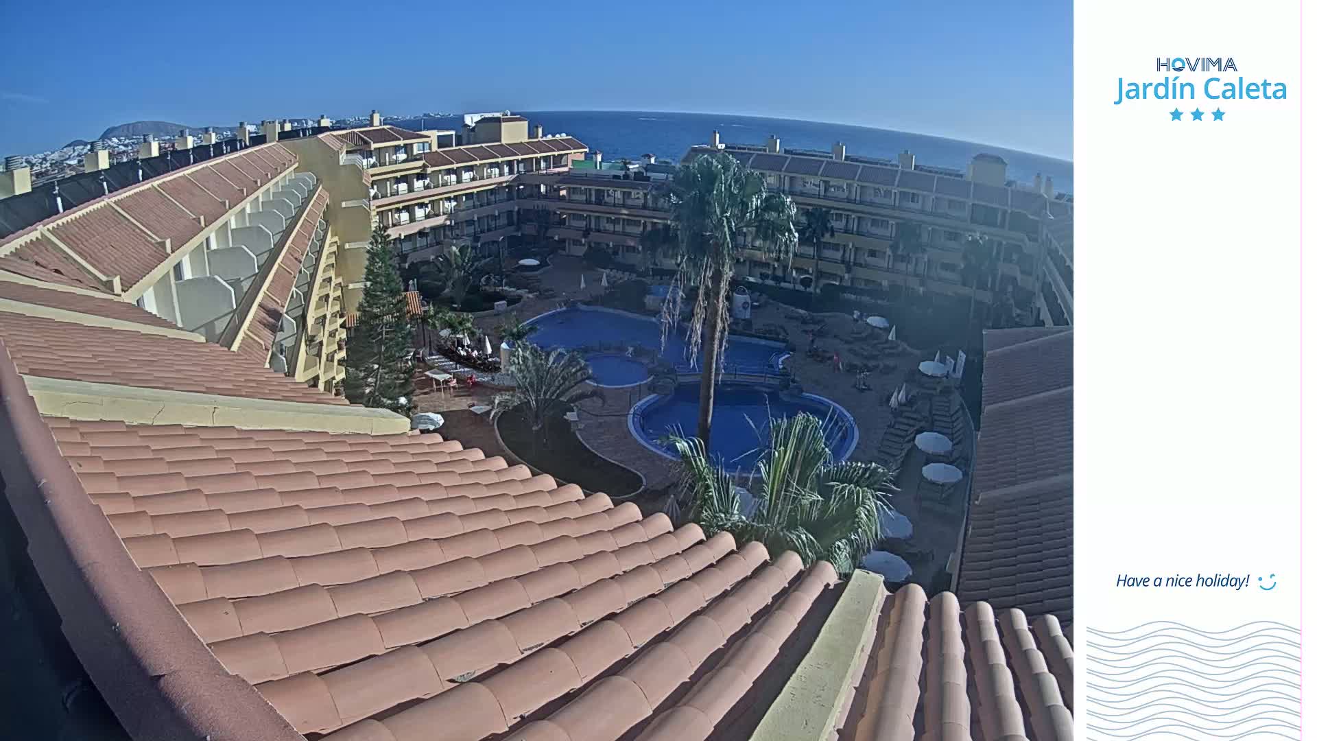 An elevated view of a resort complex with multiple pools, surrounded by buildings under a clear, sunny sky, overlooks a body of water in the distance.