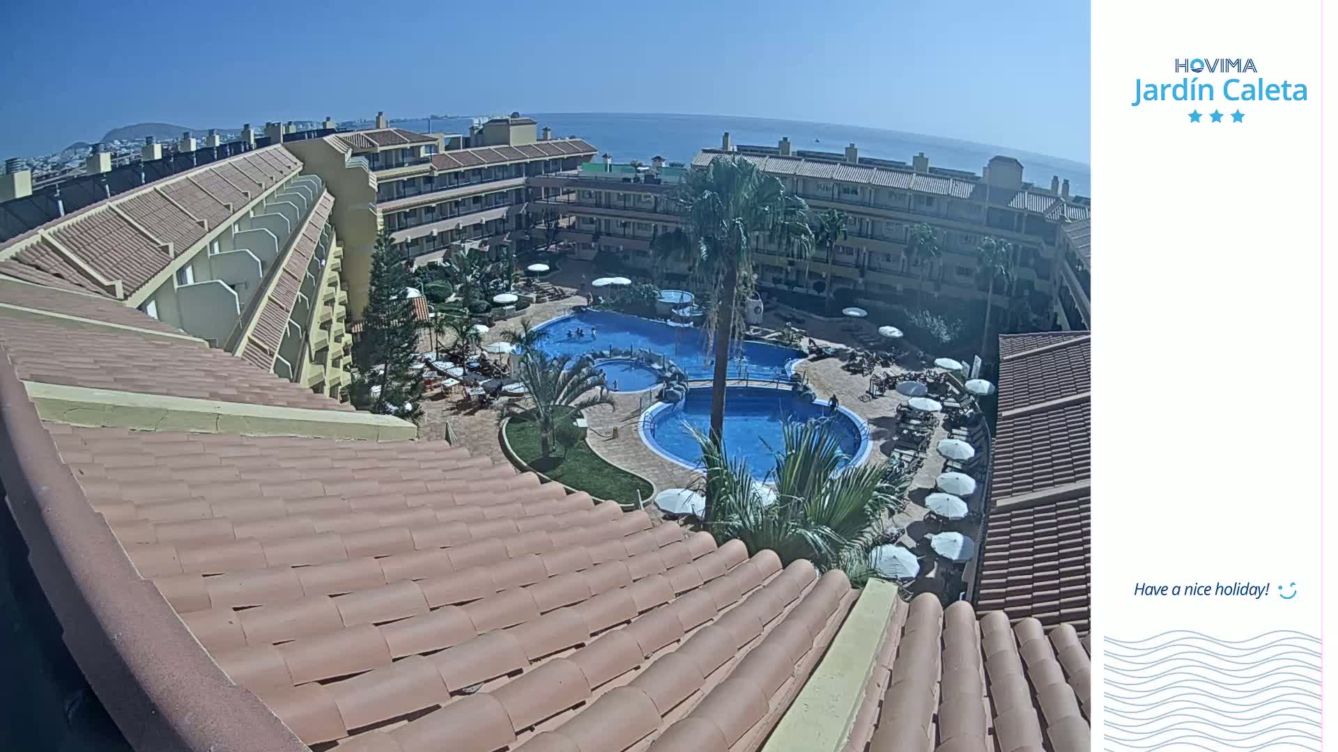 A sunny view from a rooftop shows a resort with two swimming pools surrounded by lounge chairs and palm trees, overlooking a body of water and distant mountains.