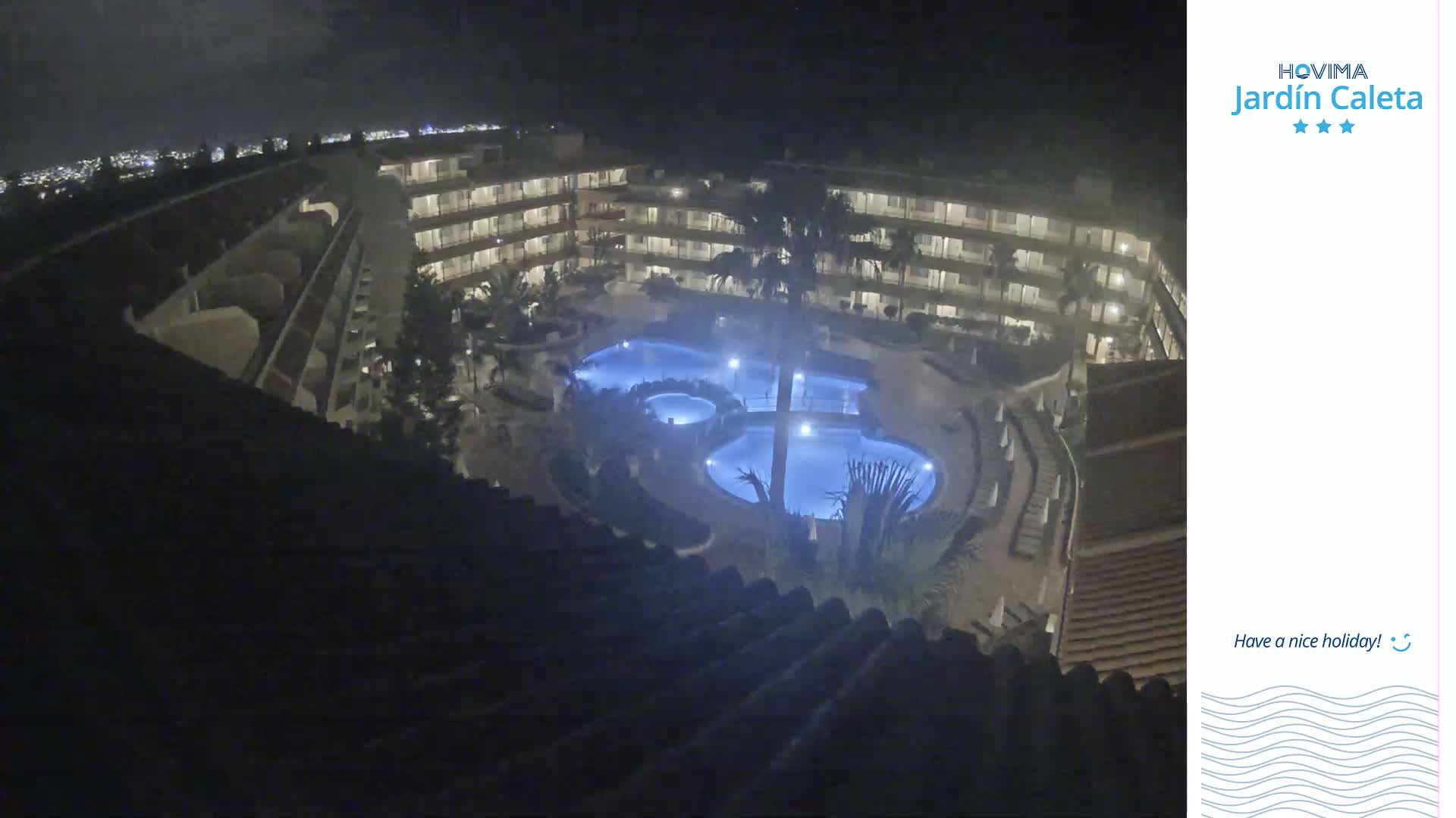 A nighttime aerial view shows a multi-story building complex surrounding illuminated swimming pools.