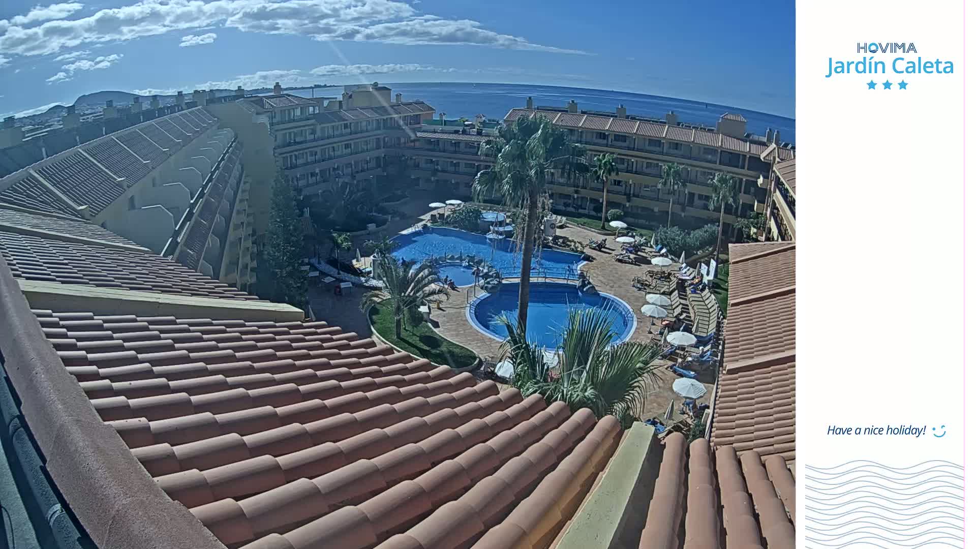 A sunny day overlooks a resort with two swimming pools surrounded by buildings and palm trees, with a view of the ocean in the distance.