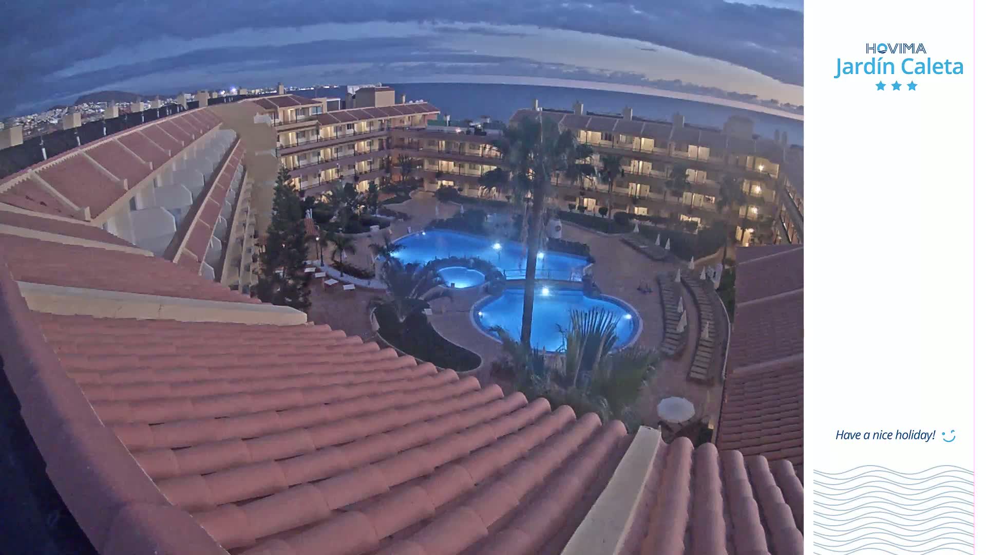 An illuminated resort pool is surrounded by low-rise buildings under a twilight sky.