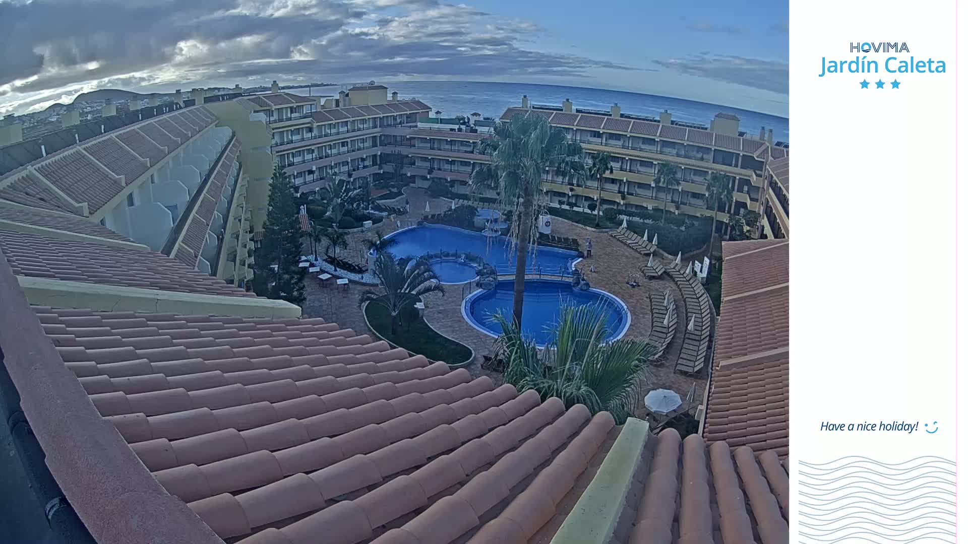 An aerial view shows a resort with two swimming pools surrounded by buildings under a partly cloudy sky.