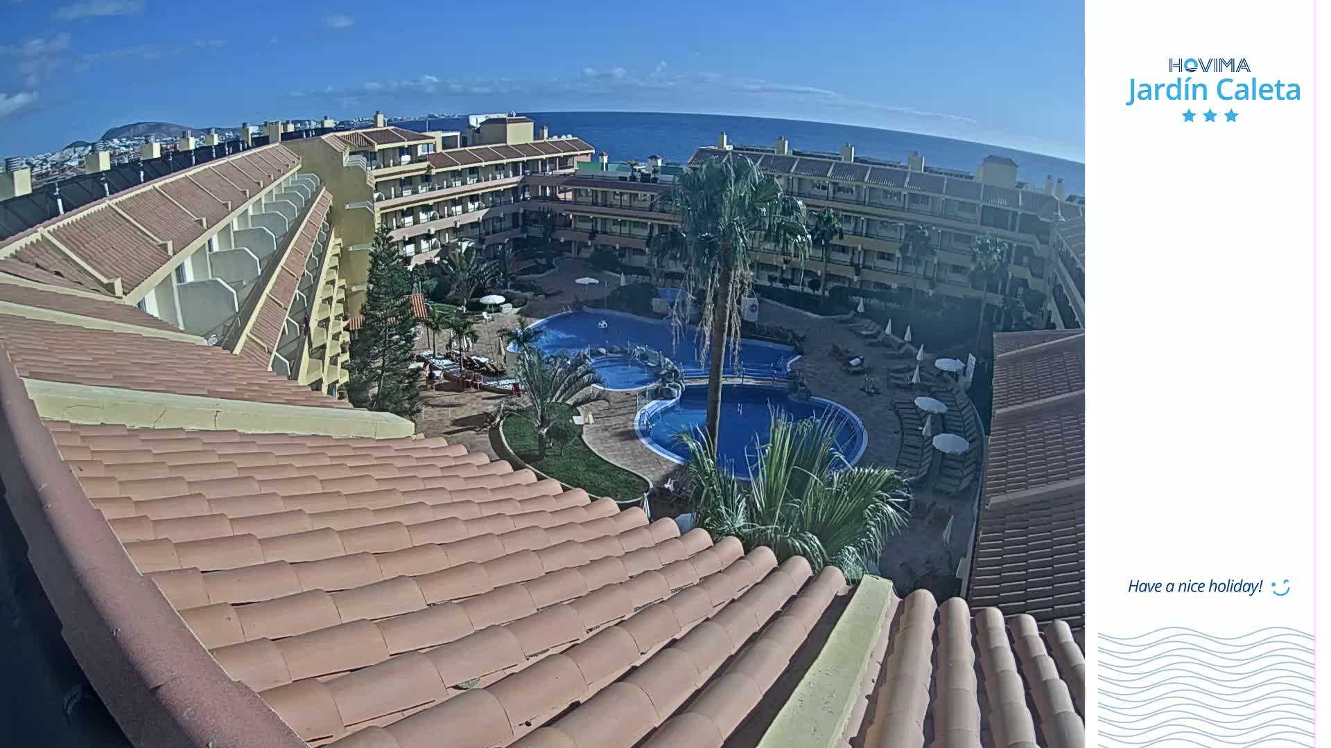 An overhead view shows a resort with multiple buildings, a large swimming pool, and palm trees, all under a clear, sunny sky with a distant ocean in the background.