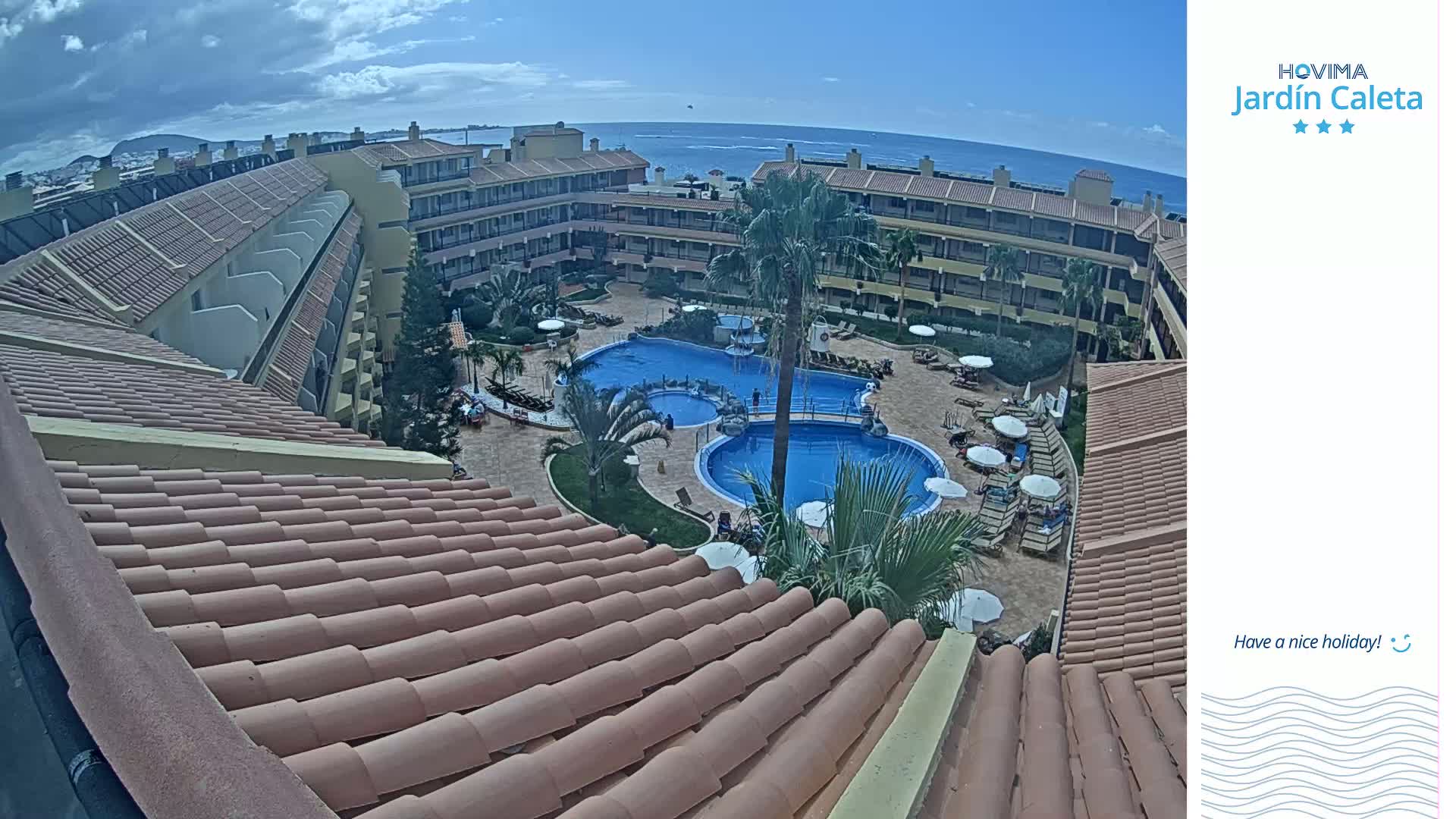 An aerial view shows a multi-story resort complex with a central courtyard containing two swimming pools, under a partly cloudy sky with the ocean visible in the distance.