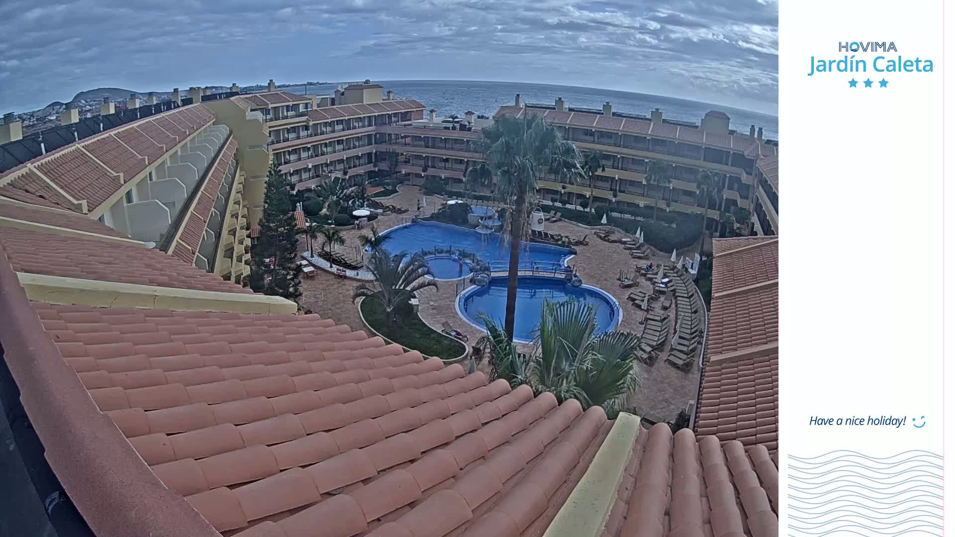An overhead view shows a resort with multiple buildings surrounding a courtyard with pools and lounge chairs under a partly cloudy sky.