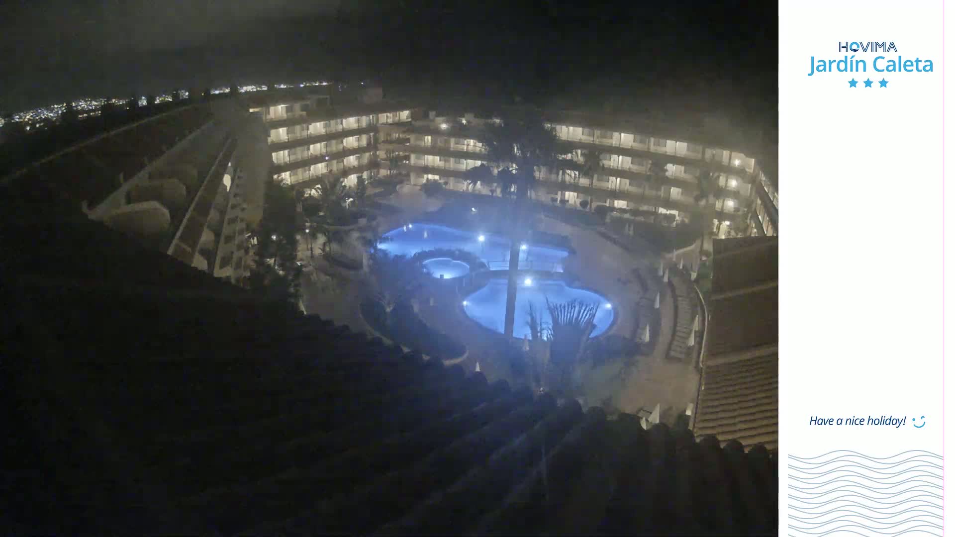 A nighttime, aerial view shows a multi-story building complex surrounding a lit, kidney-shaped swimming pool.