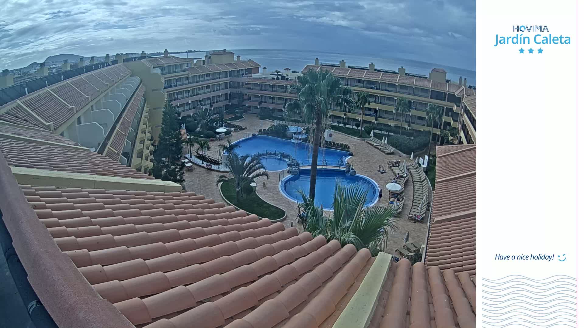 A view from a rooftop shows a resort courtyard with multiple pools surrounded by buildings under a cloudy sky.