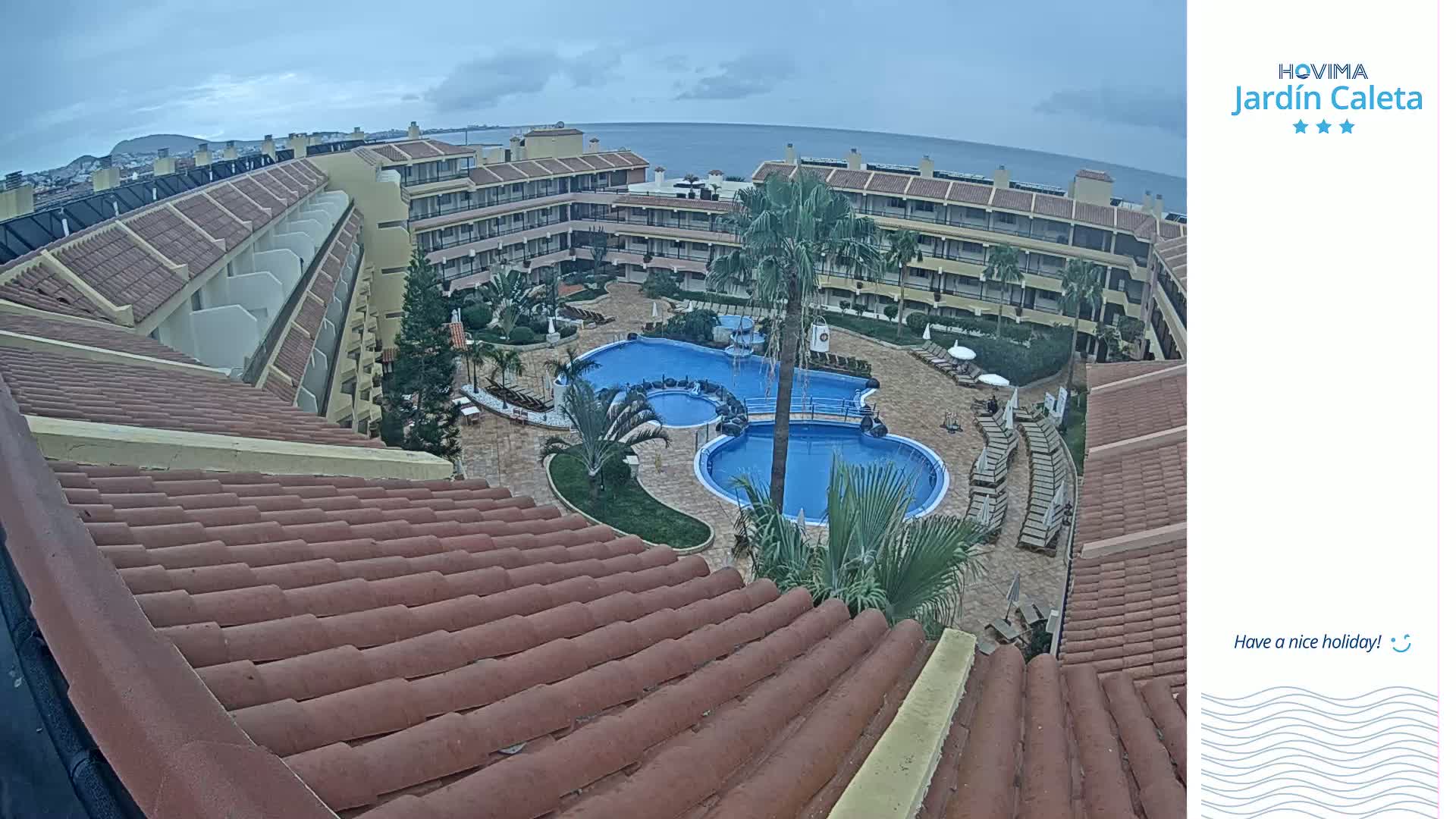 An overhead view of a resort pool area on an overcast day, showing two pools surrounded by buildings and lounge chairs, overlooking the ocean.
