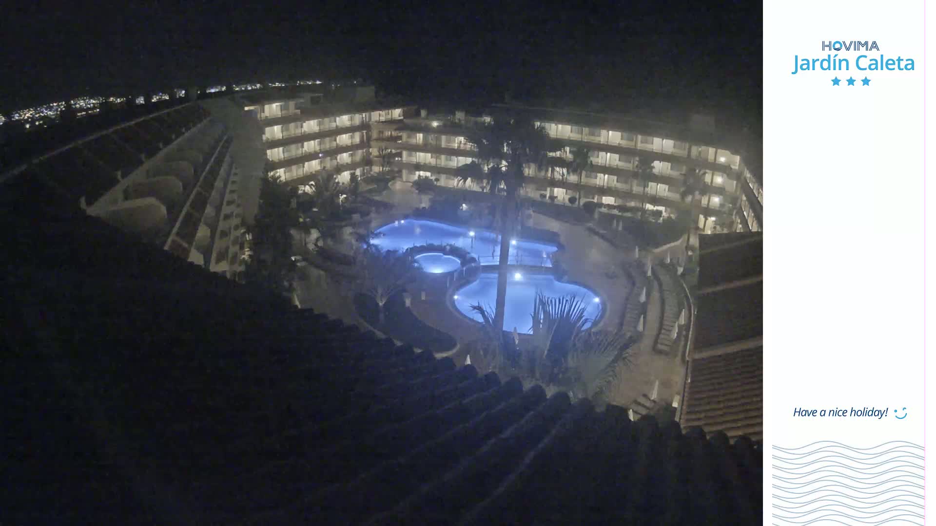 An illuminated resort pool and surrounding buildings are viewed at night from an elevated vantage point.