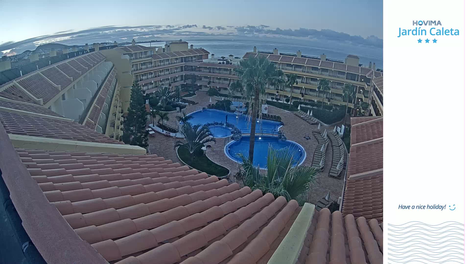 A tiled roof overlooks a courtyard with a swimming pool and surrounding apartment buildings under a partly cloudy sky.
