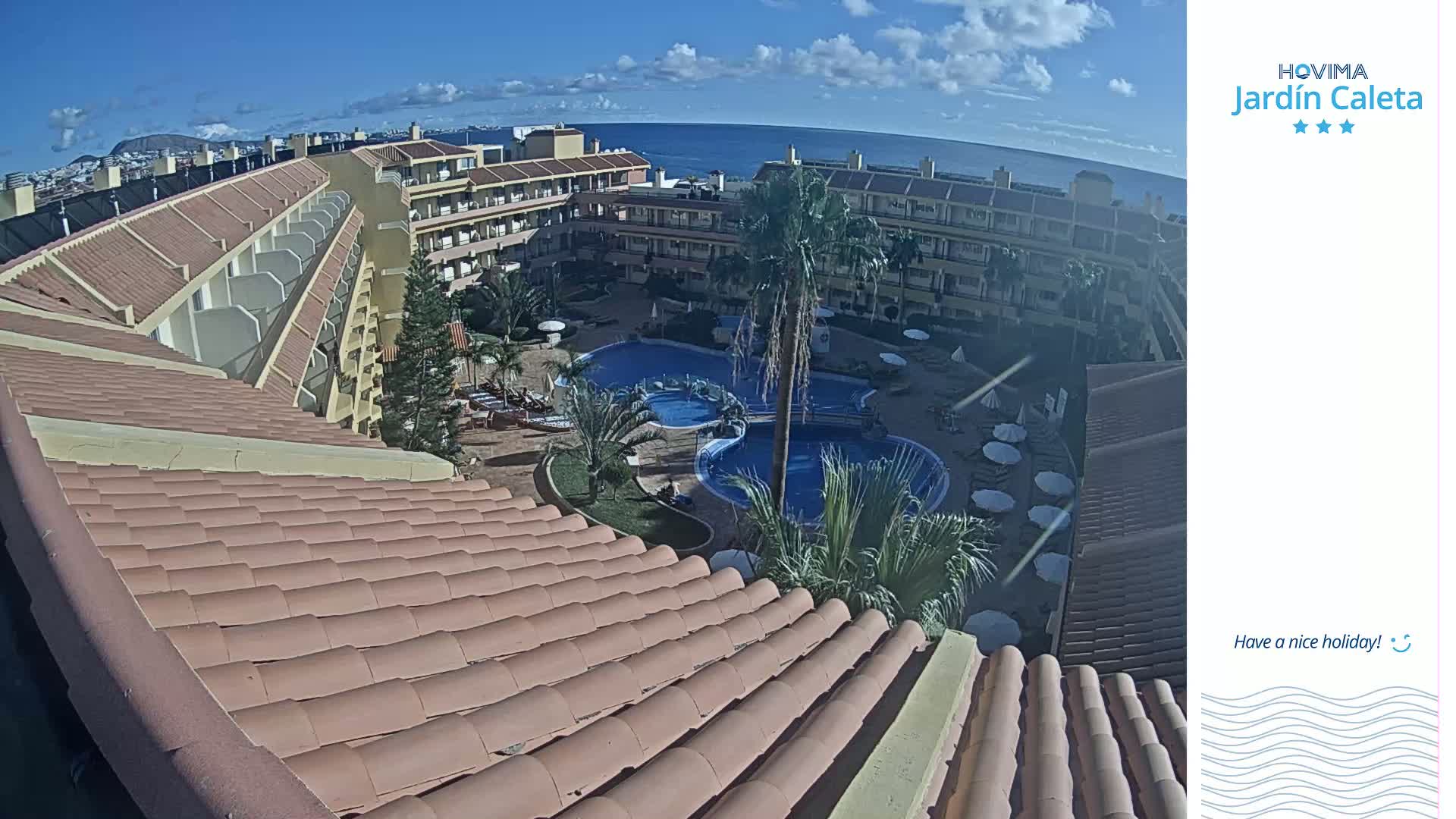 A curved tiled roof overlooks a courtyard with two swimming pools, palm trees, and lounge chairs, all under a sunny sky and with a distant ocean view.
