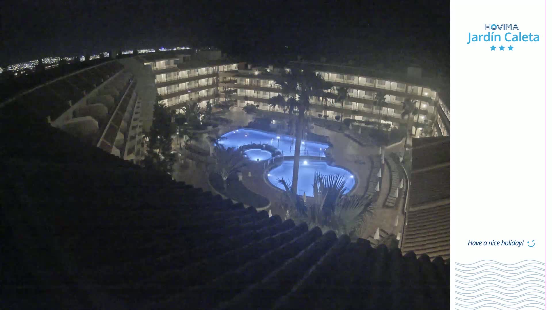 A nighttime, overhead view of a multi-story building complex with multiple illuminated swimming pools, palm trees, and surrounding landscaping.