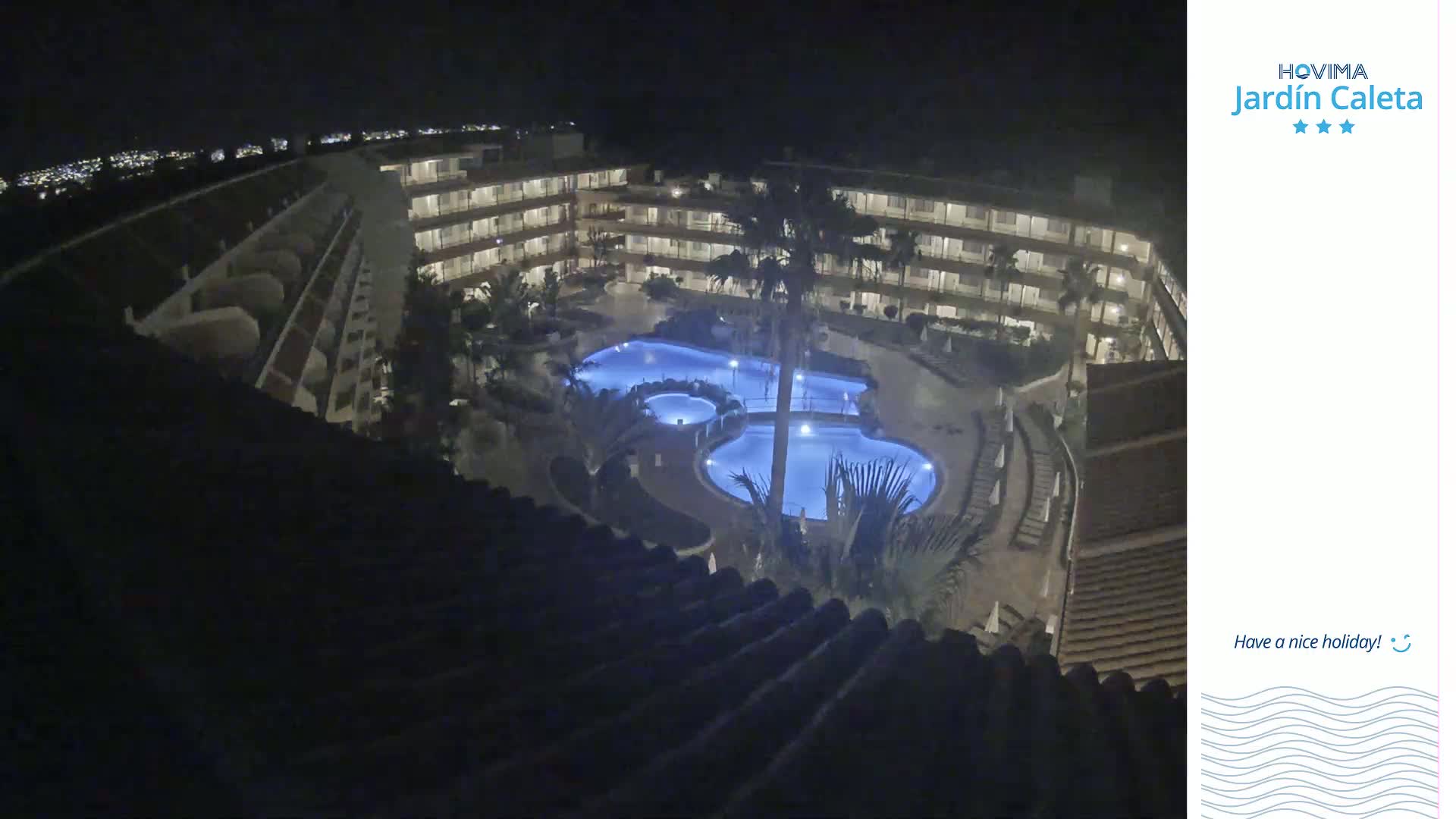 A nighttime, overhead view shows a multi-story building complex surrounding a lit, kidney-shaped swimming pool.