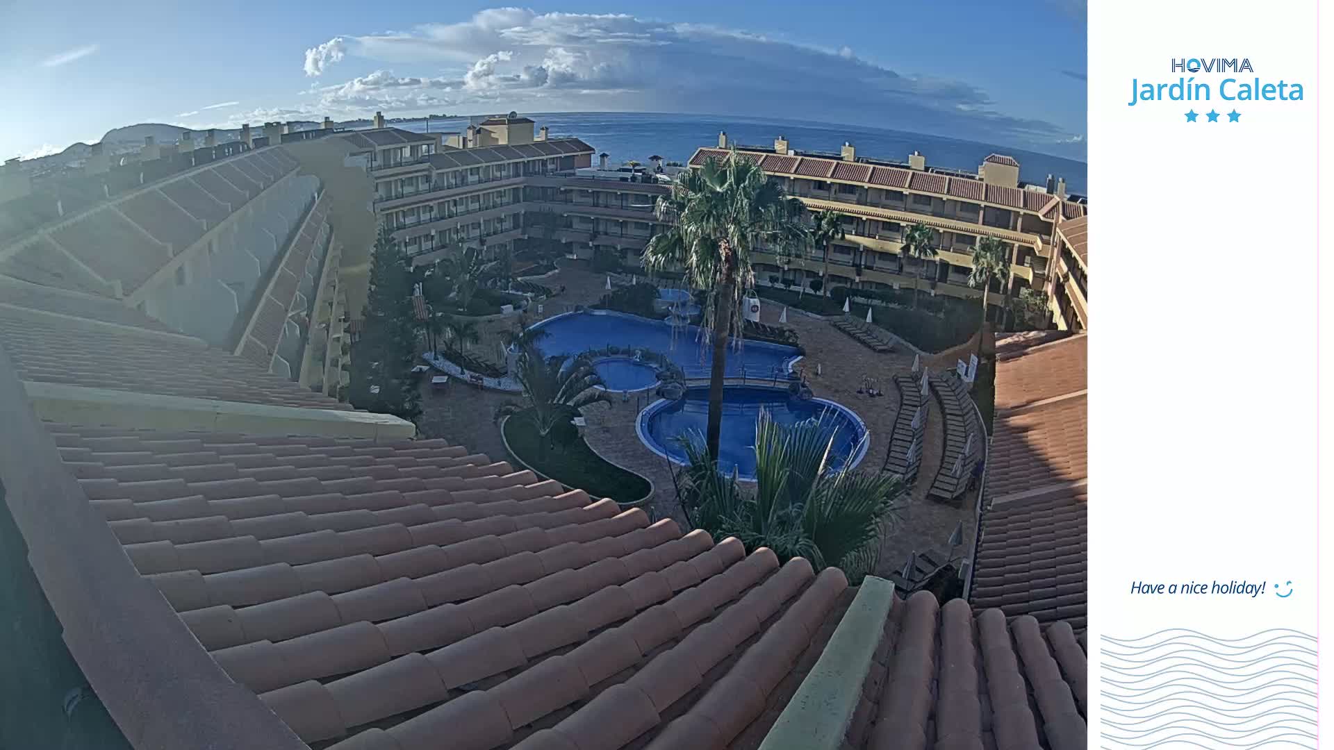An aerial view shows a resort with a swimming pool complex under partly cloudy skies.