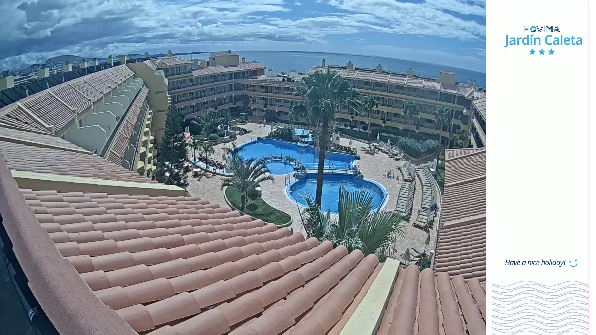 An elevated view of a resort complex with multiple swimming pools, palm trees, and sun loungers, under a partly cloudy sky.
