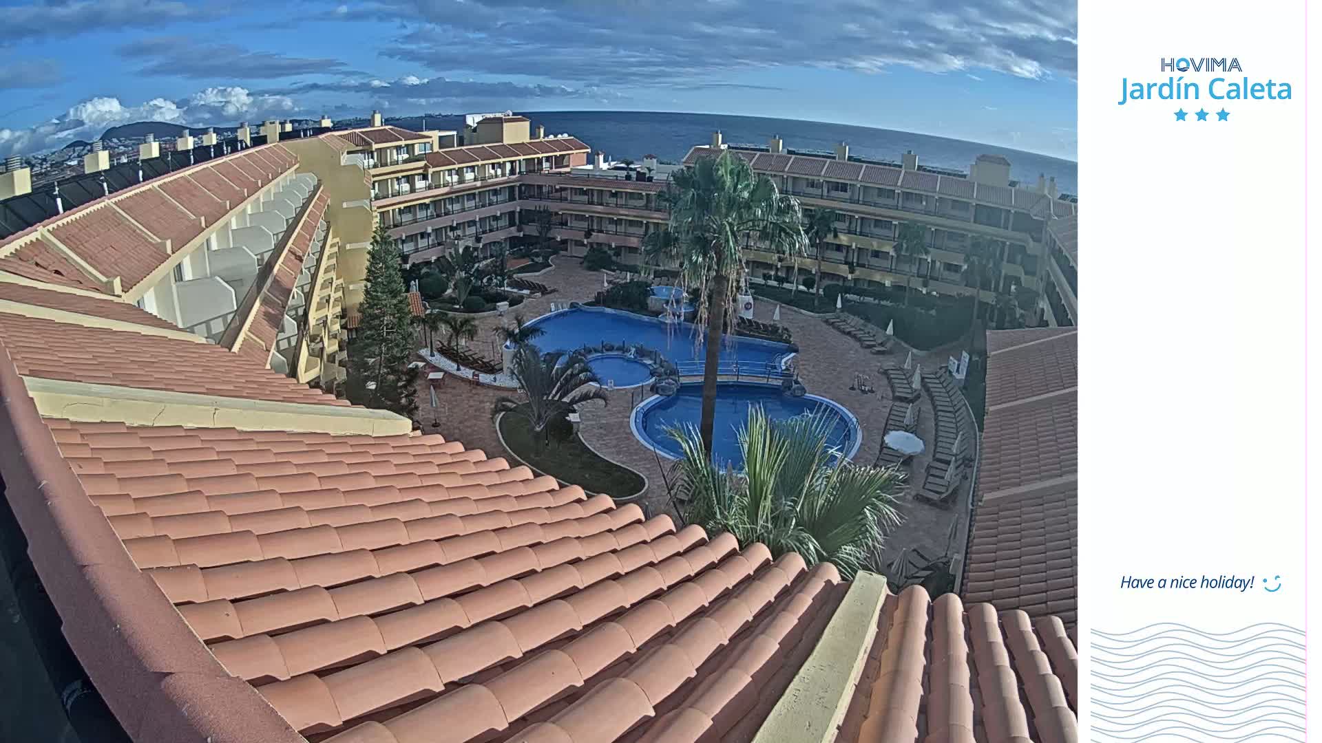 An overhead view of a resort complex with multiple buildings, a central swimming pool area, and a palm tree, under a partly cloudy sky.