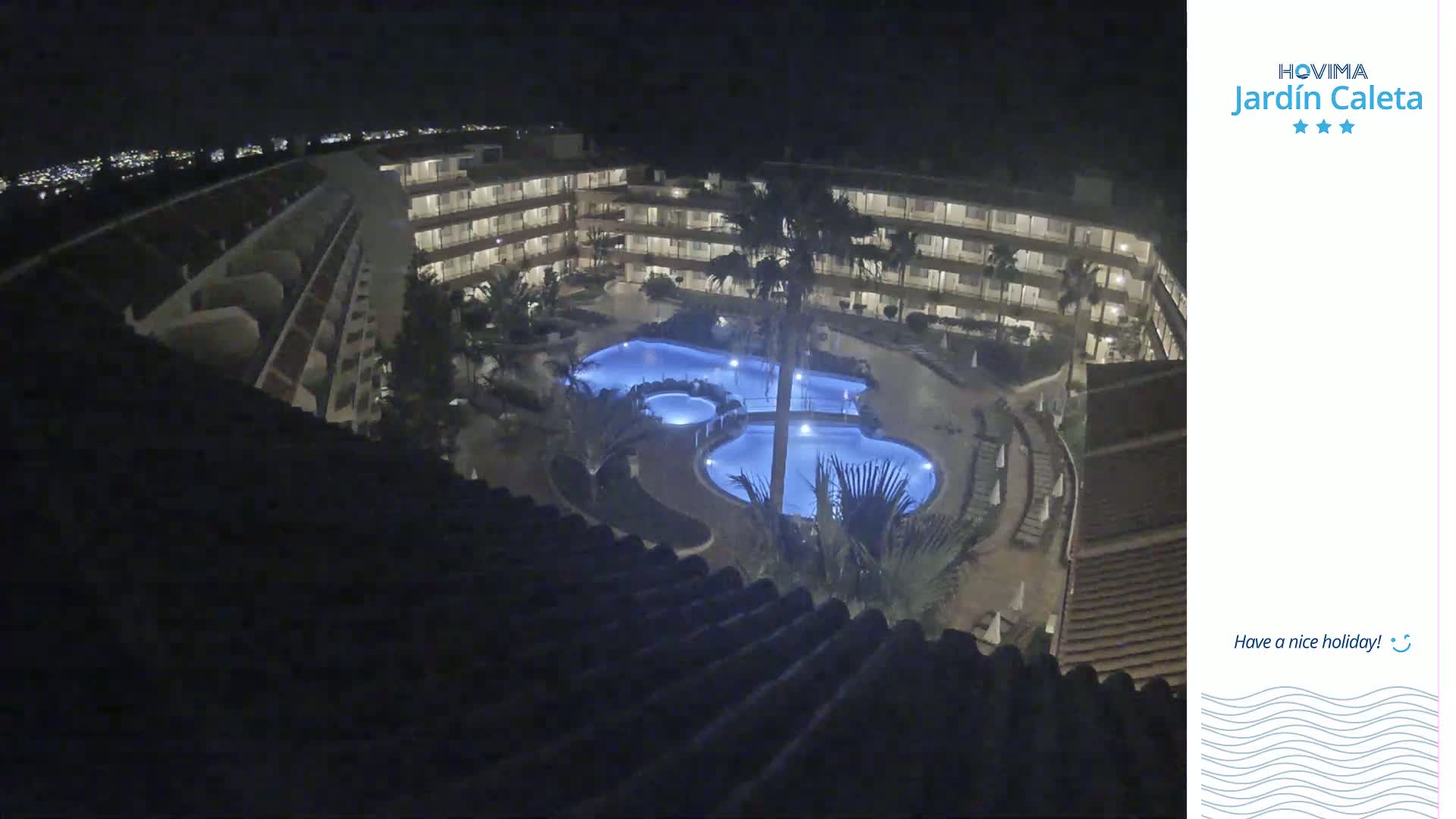A nighttime aerial view shows a multi-story hotel complex with a large, illuminated swimming pool surrounded by landscaping.