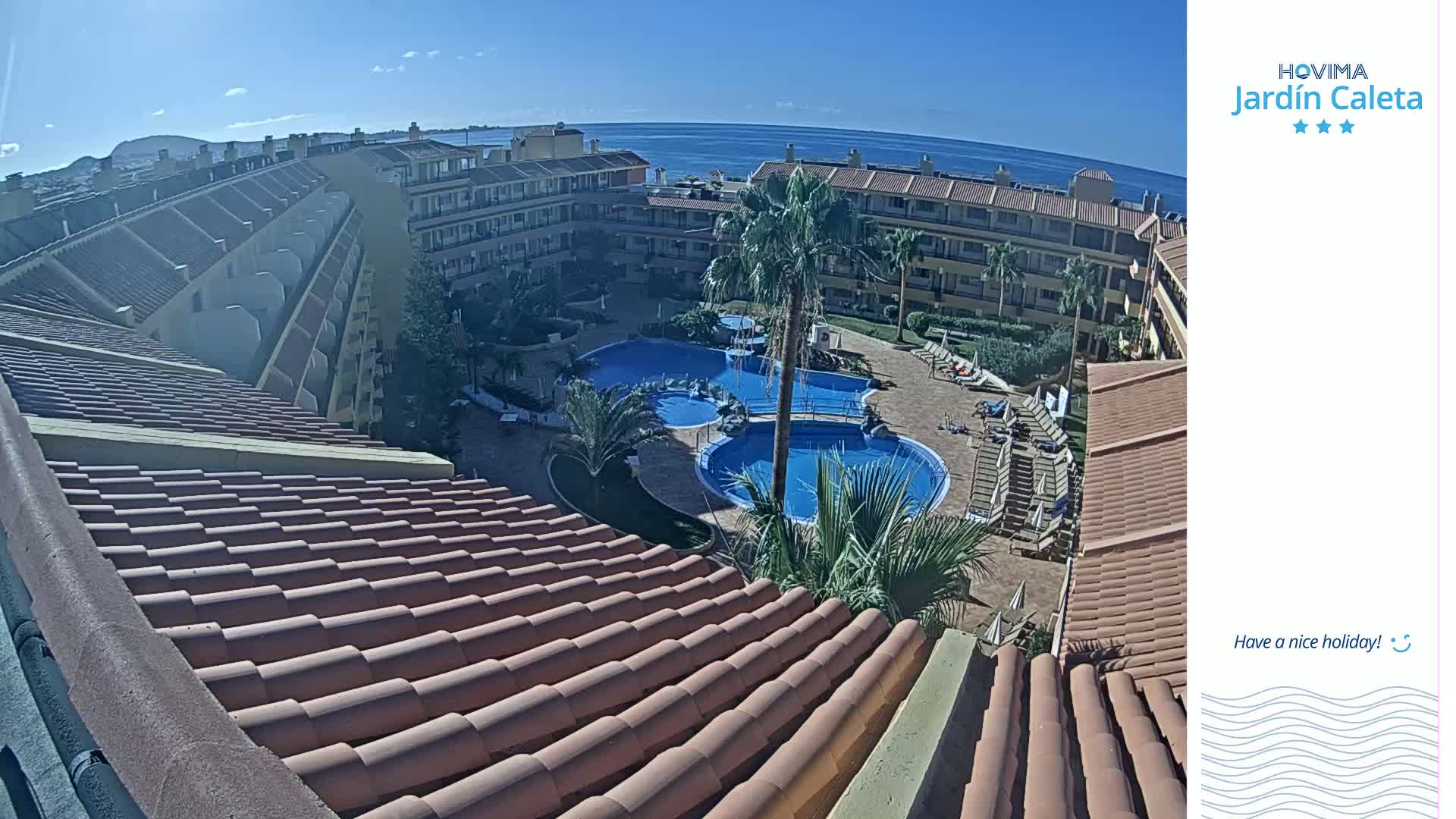 A sunny view from a rooftop shows a courtyard with a swimming pool and surrounding apartment buildings overlooking the ocean.