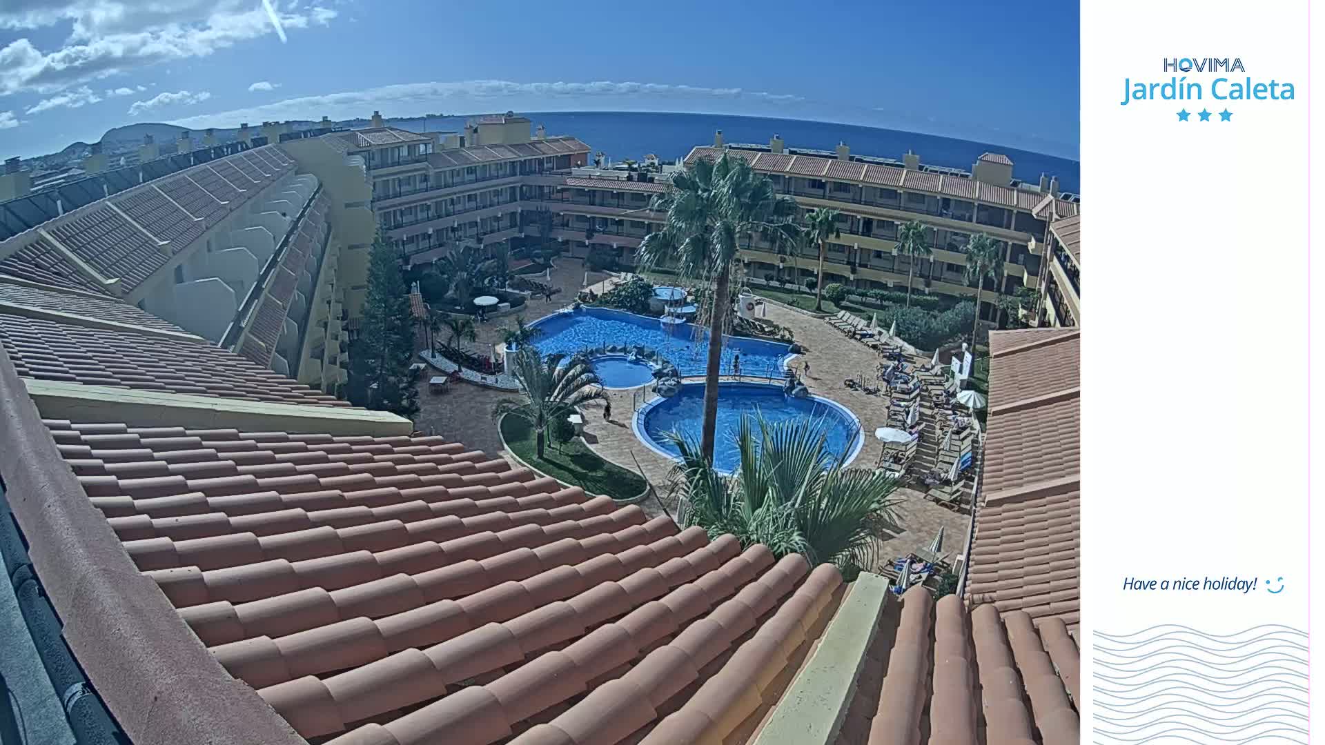 A sunny day overlooks a resort with multiple pools surrounded by buildings, viewed from a tiled rooftop.