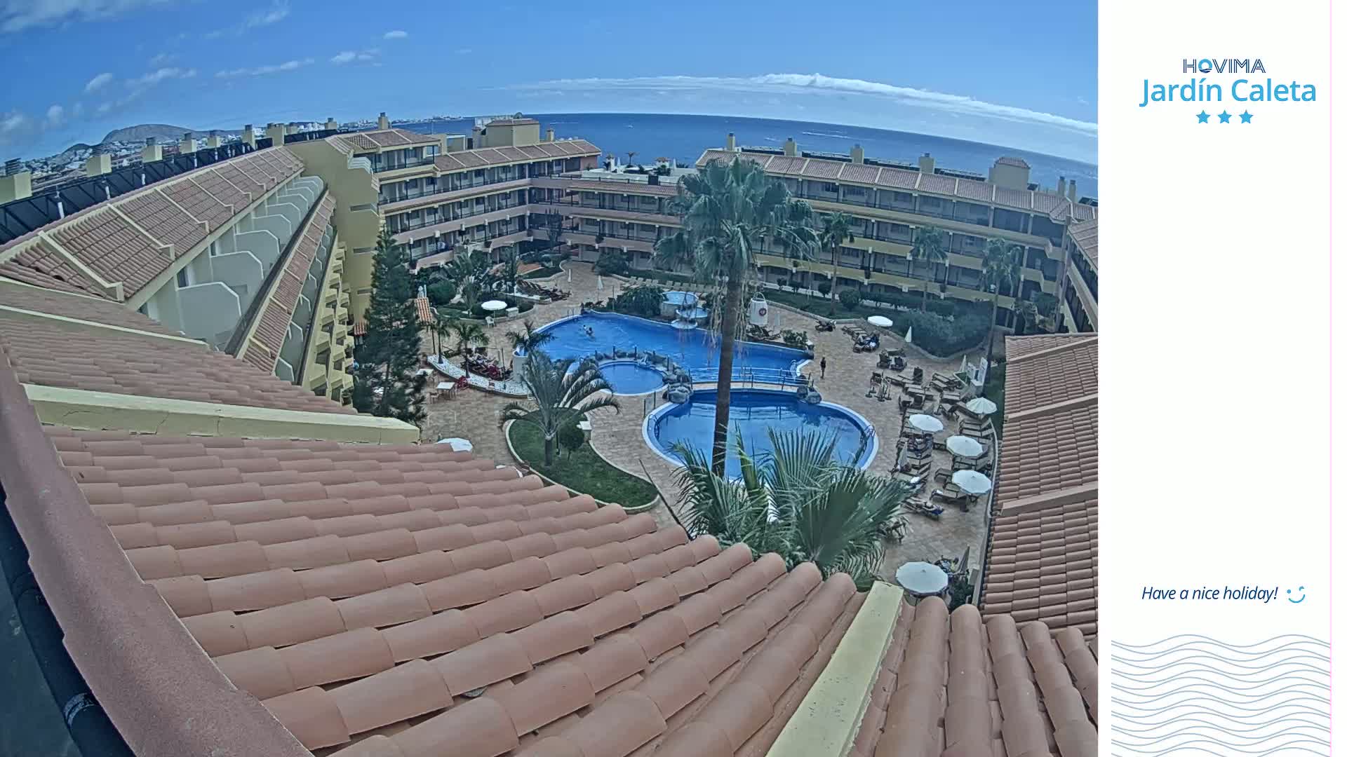 A tiled roof overlooks a resort courtyard with two swimming pools, palm trees, and lounge chairs under a mostly sunny sky.