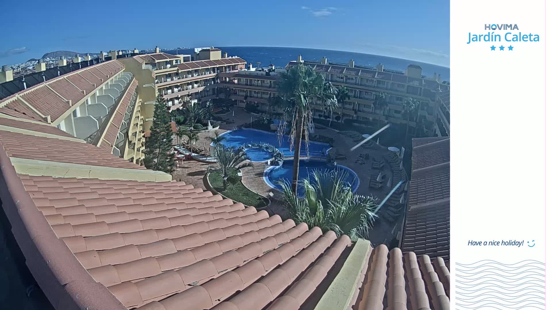 A tiled roof overlooks a courtyard with swimming pools and palm trees, all under a clear, sunny sky and a distant ocean view.
