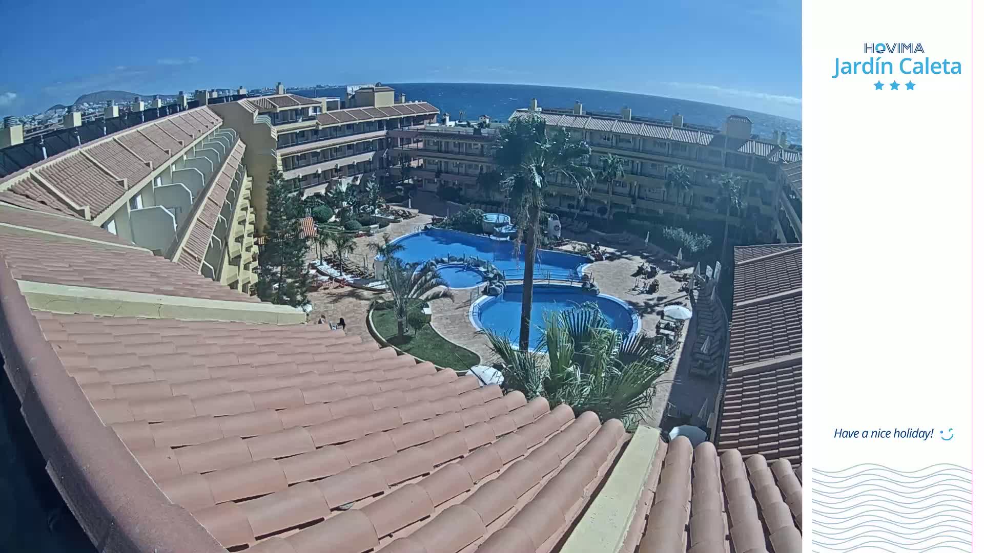 A sunny view from a rooftop shows a resort complex with multiple swimming pools and ocean in the background.