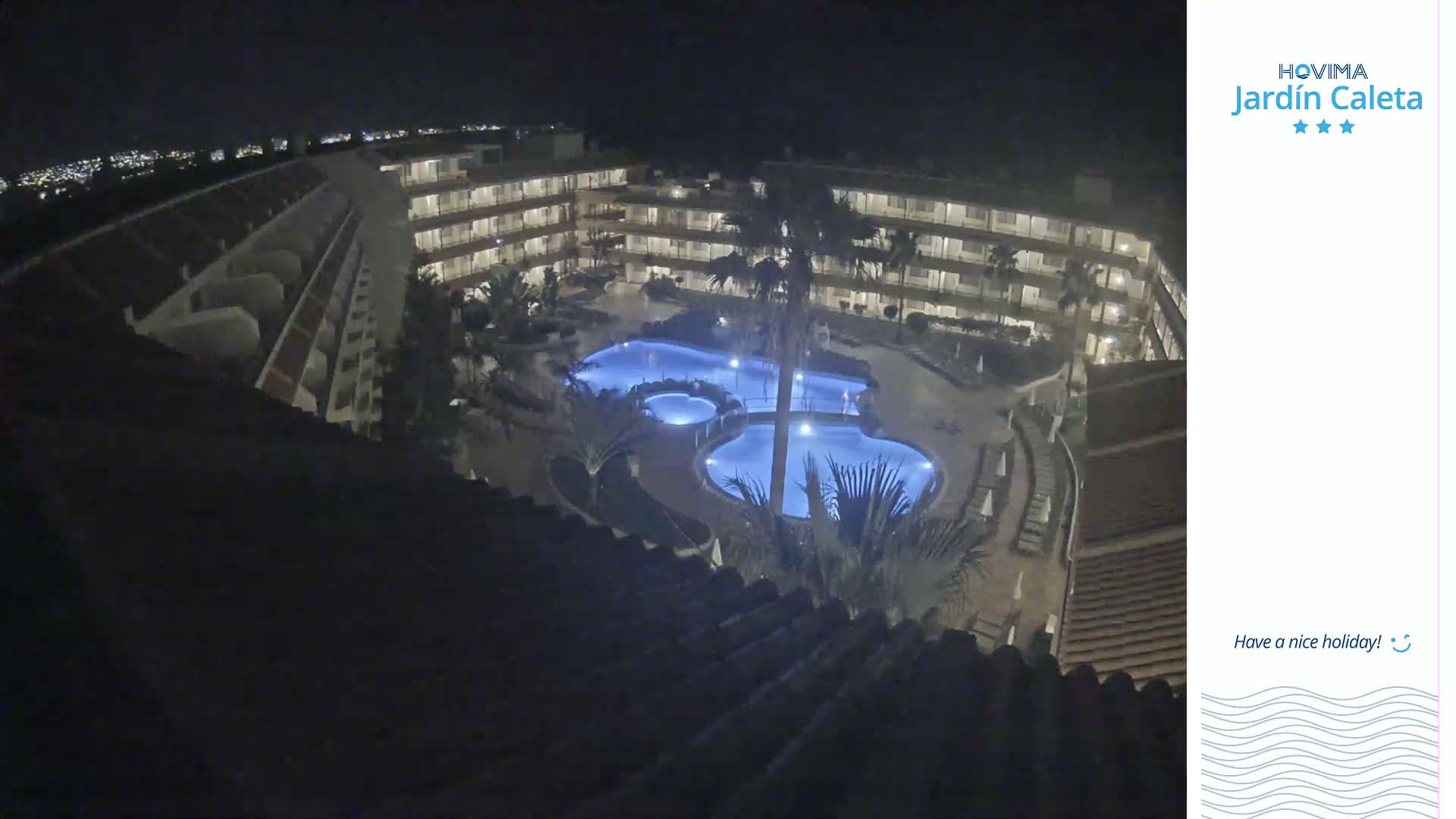 A nighttime aerial view shows a multi-story building complex surrounding a lit-up, irregularly shaped swimming pool.