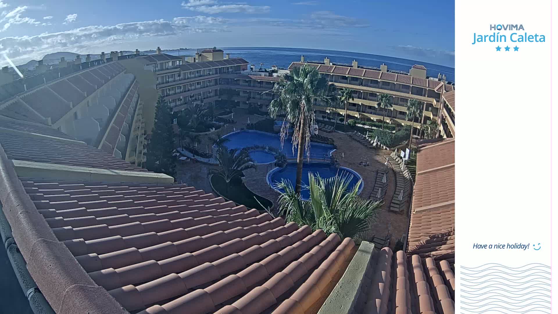 An overhead view shows a resort with a swimming pool complex under a partly cloudy sky, overlooking the ocean.