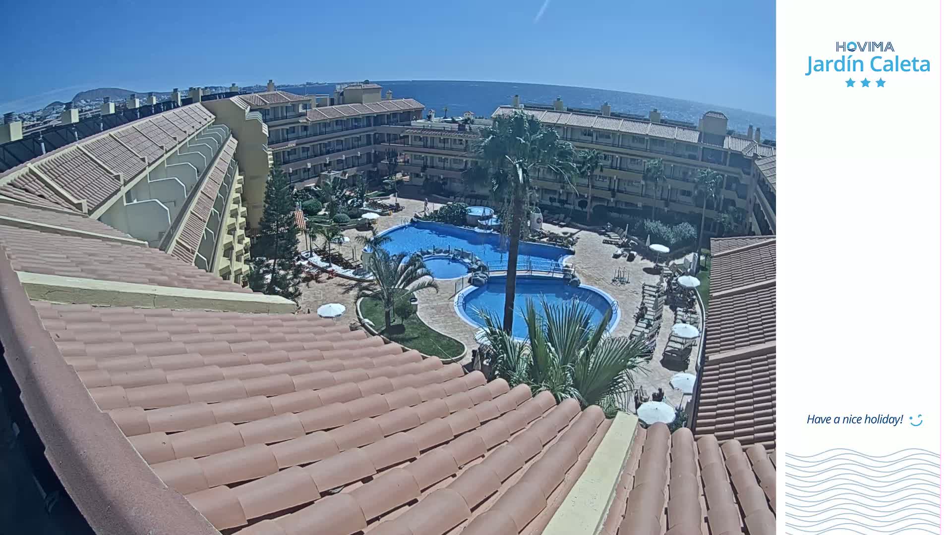 A sunny view from a rooftop shows a resort with multiple pools surrounded by tan buildings and palm trees, overlooking a calm ocean.