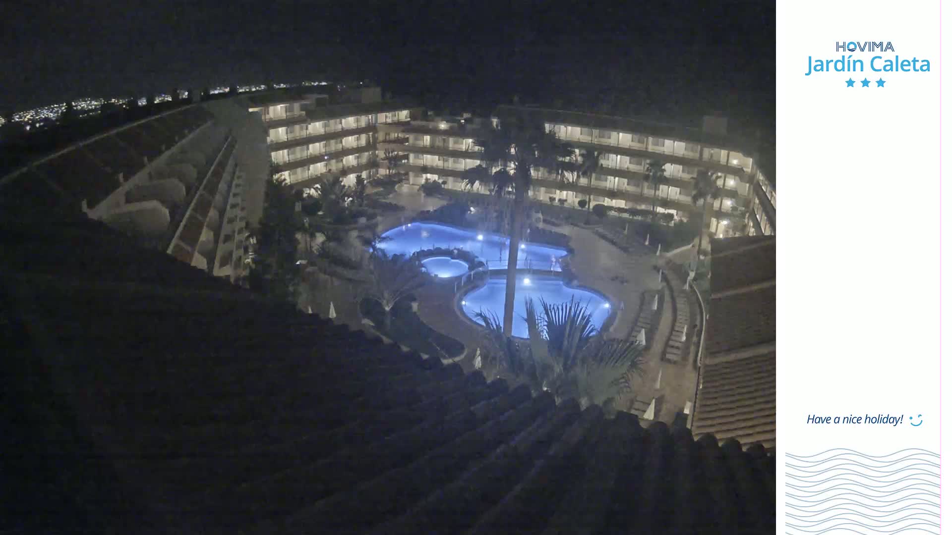 A nighttime, high-angle view shows a multi-story hotel complex with a swimming pool illuminated in blue.