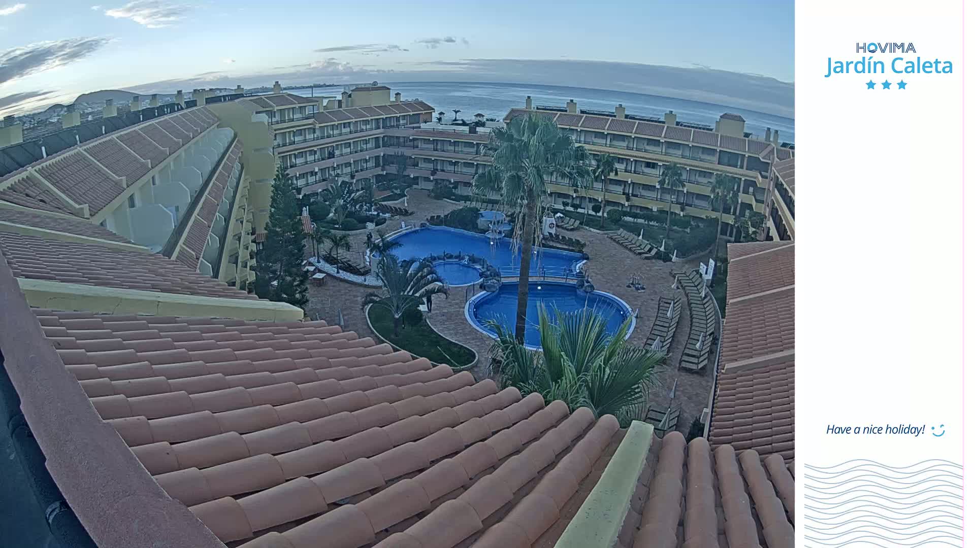 A tiled roof overlooks a courtyard with a swimming pool and a low-rise building complex, under a clear, twilight sky.