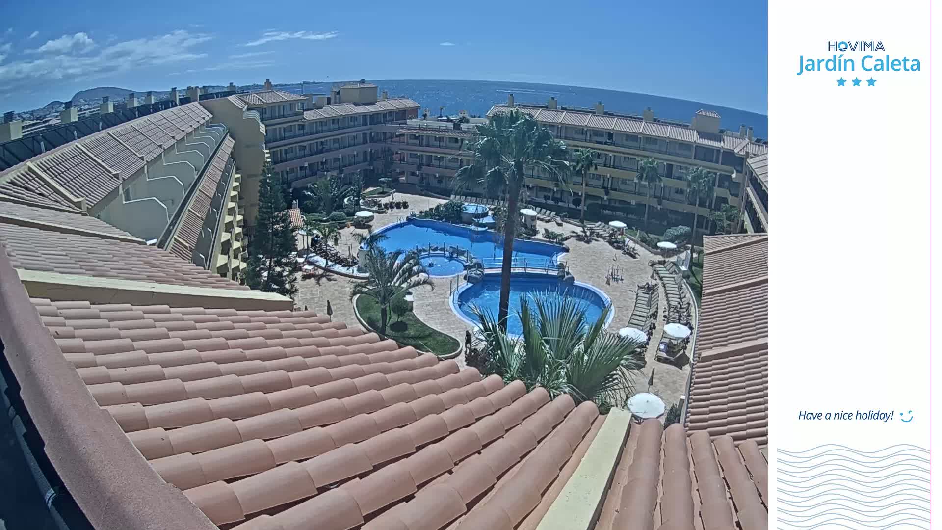 An aerial view shows a resort with multiple buildings surrounding a pool area on a sunny day, overlooking the ocean.