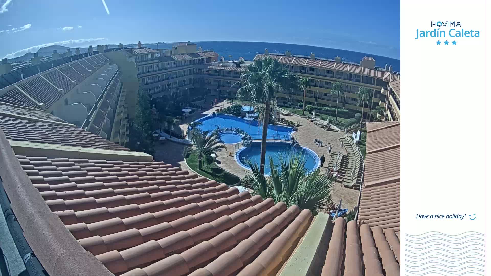 A rooftop view overlooking a sunny courtyard with two swimming pools, surrounded by a low-rise apartment complex and palm trees, next to a calm ocean.
