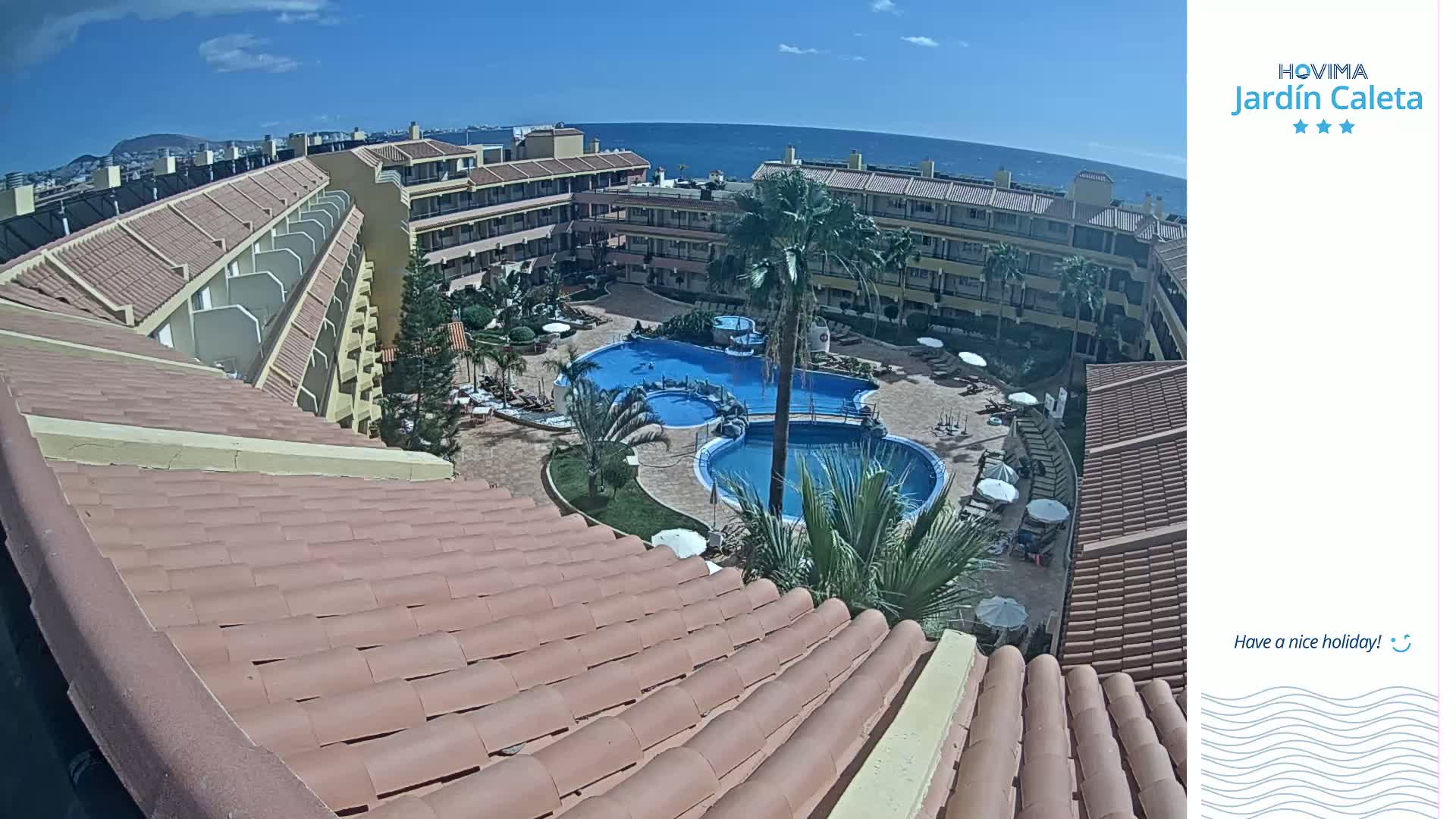 An aerial view of a resort complex with multiple pools, surrounded by buildings and palm trees, on a sunny day overlooking the ocean.