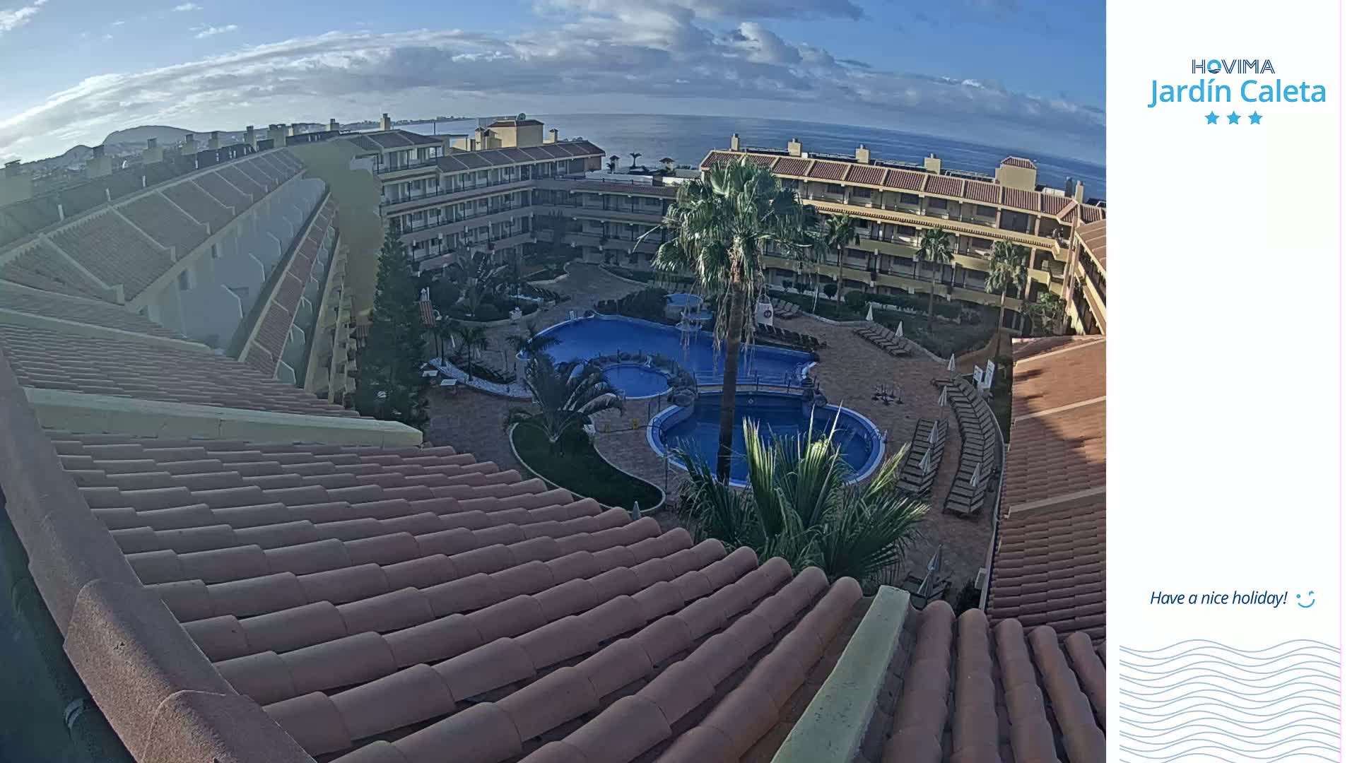 An aerial view shows a resort with a multi-level building surrounding a central pool area under a partly cloudy sky.