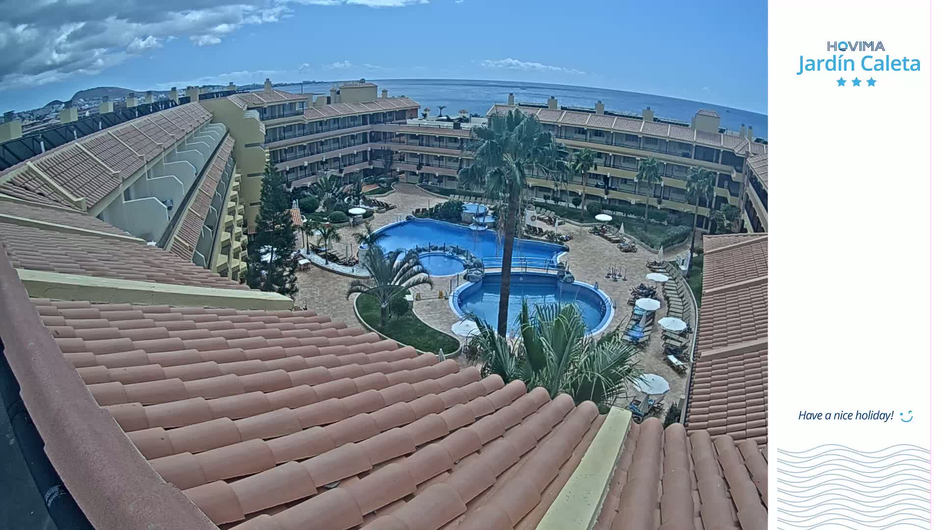 An overhead view shows a resort courtyard with two swimming pools surrounded by a building with a reddish-brown tile roof, under partly cloudy skies and overlooking a calm ocean.