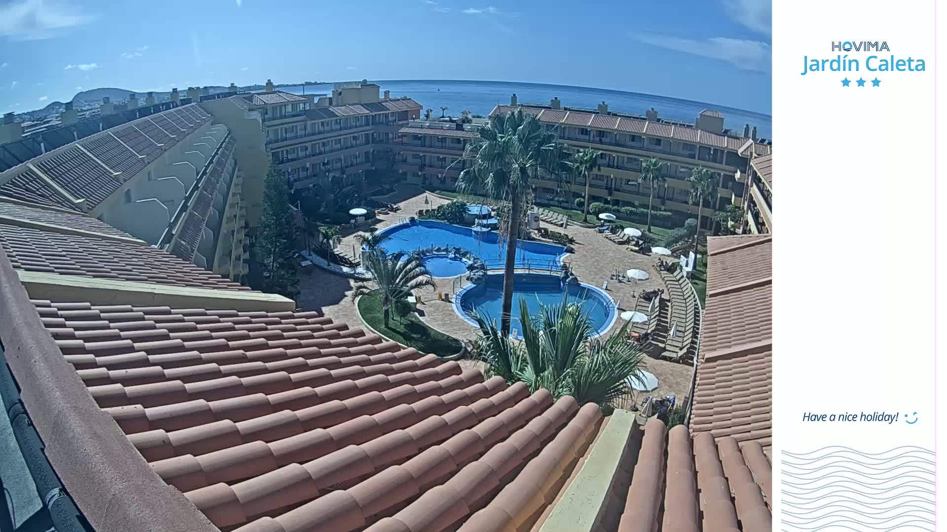An aerial view of a resort complex with multiple swimming pools, surrounded by buildings and palm trees under sunny skies.