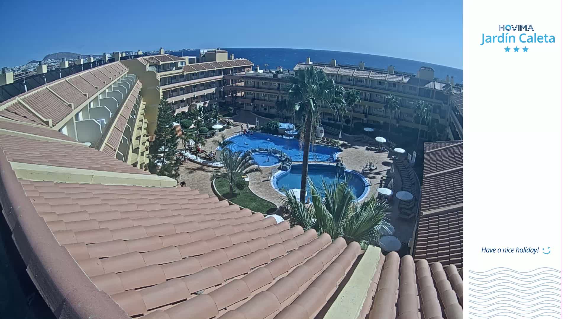 A sun-drenched view from a rooftop shows a resort complex with multiple pools, palm trees, and a clear blue ocean in the distance.