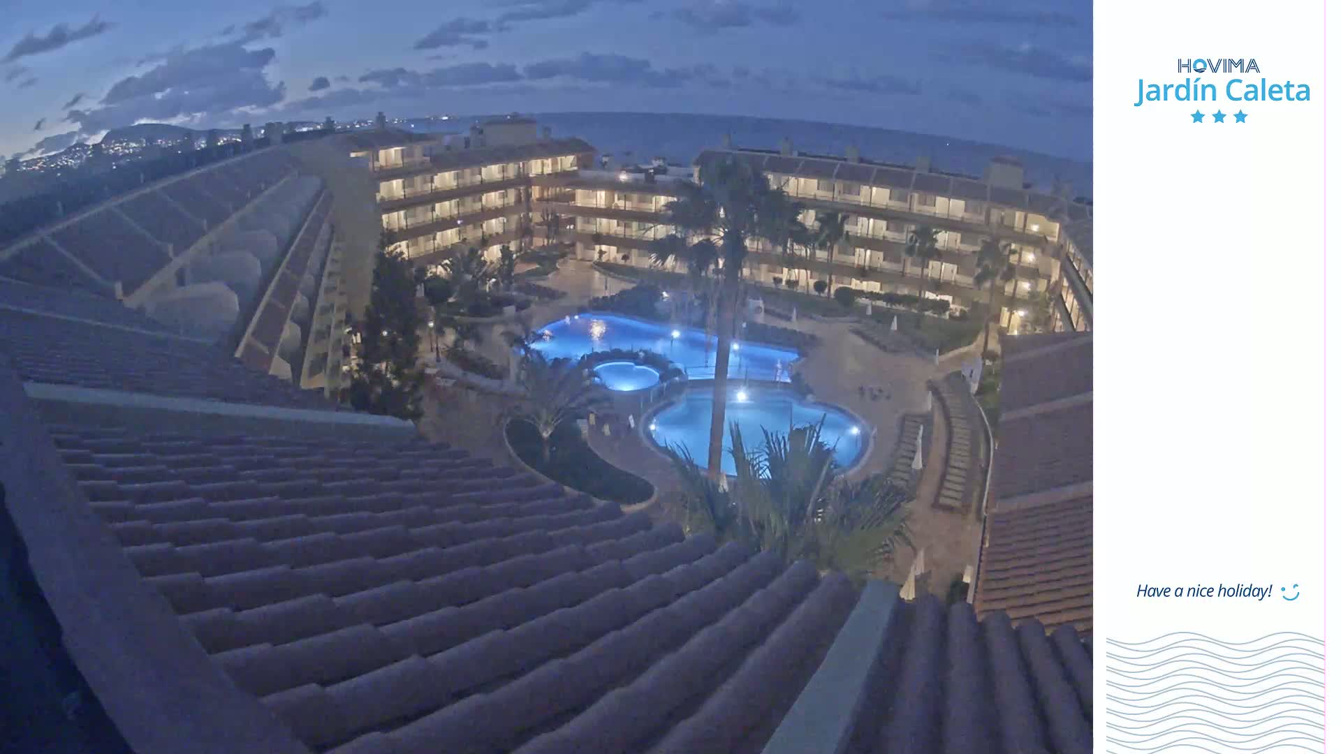 An illuminated apartment complex with multiple swimming pools is seen at night under a dark blue sky.
