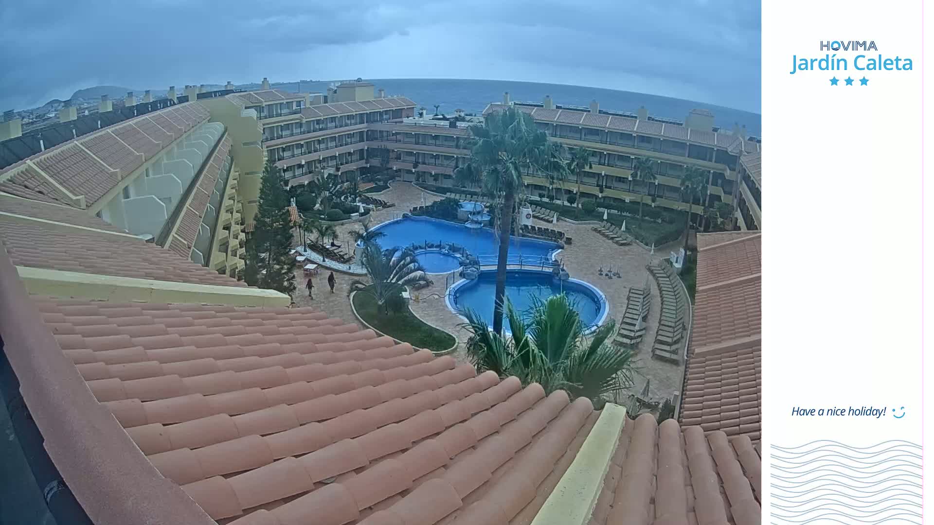 An overhead view of a resort's courtyard, featuring a multi-sectioned pool surrounded by lounge chairs and buildings under an overcast sky.