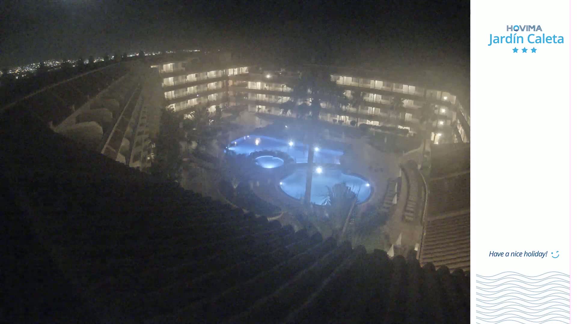 A nighttime, hazy view from above shows a multi-story building complex surrounding a lit swimming pool.