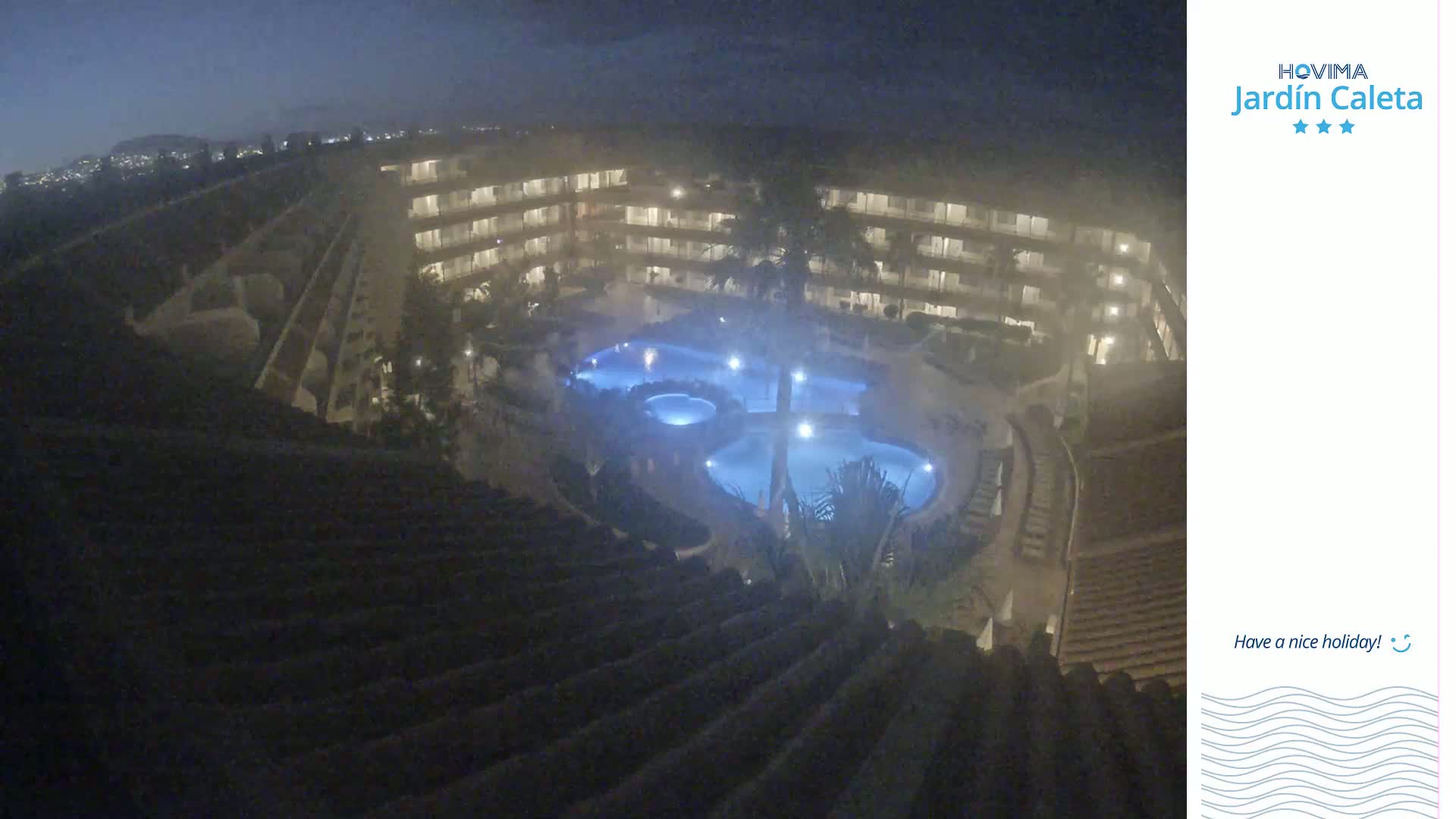 A nighttime, high-angle view shows a multi-story building complex surrounding a lit, multi-sectioned swimming pool.