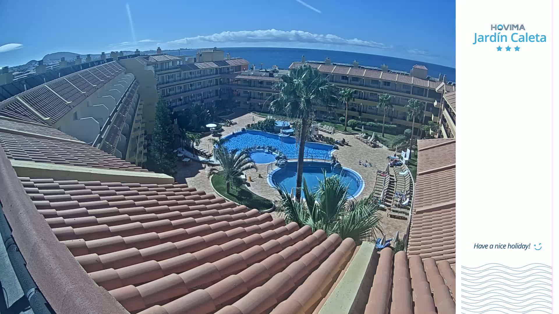 A sunny day overlooks a resort pool area with several buildings and tiled roofs, a palm tree, and a clear view of the ocean in the distance.
